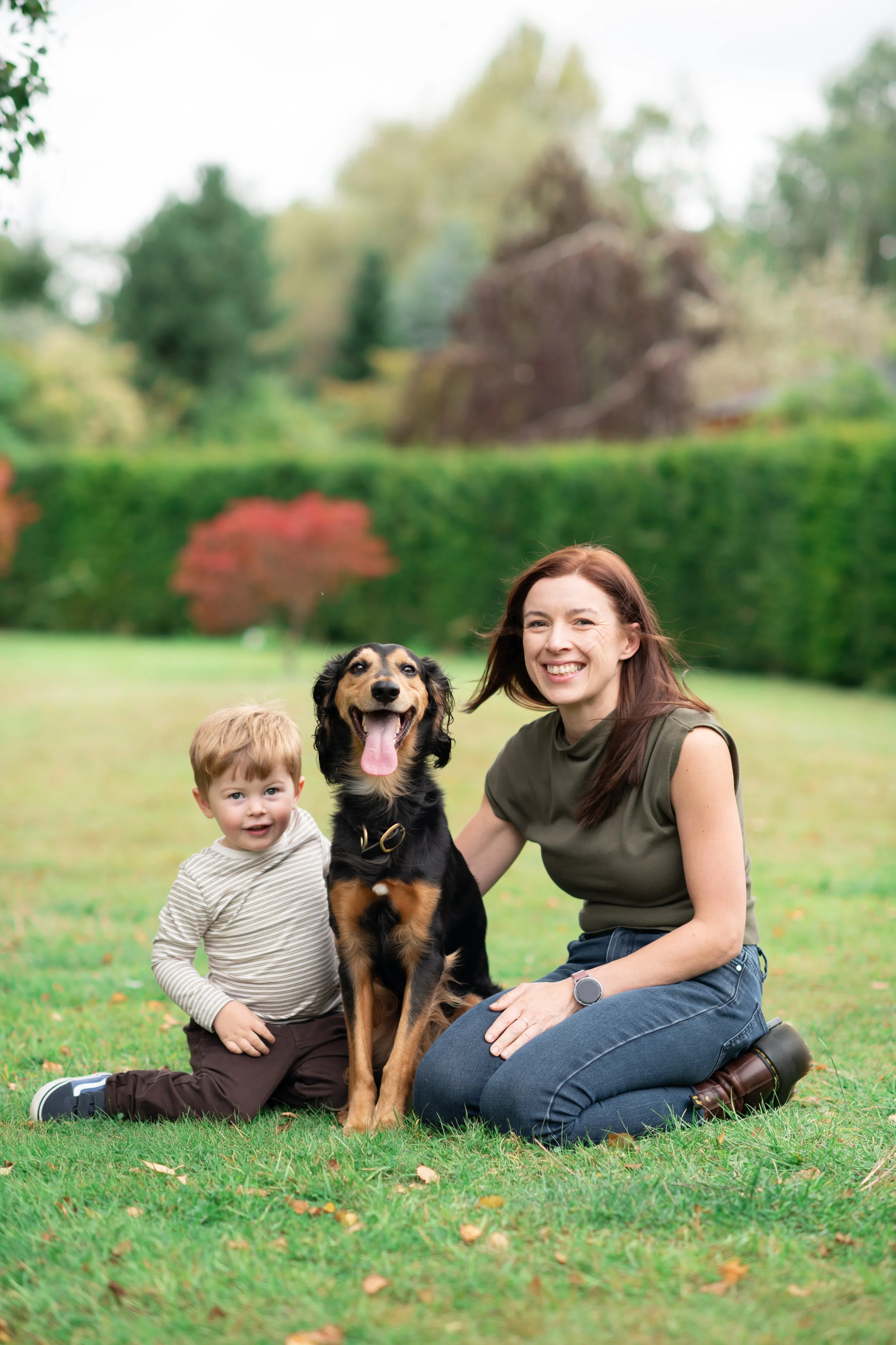 A woman, a young boy, and a black and tan dog sitting on a grassy lawn outdoors with trees in the background. The woman and dog are smiling, and the boy appears happy.