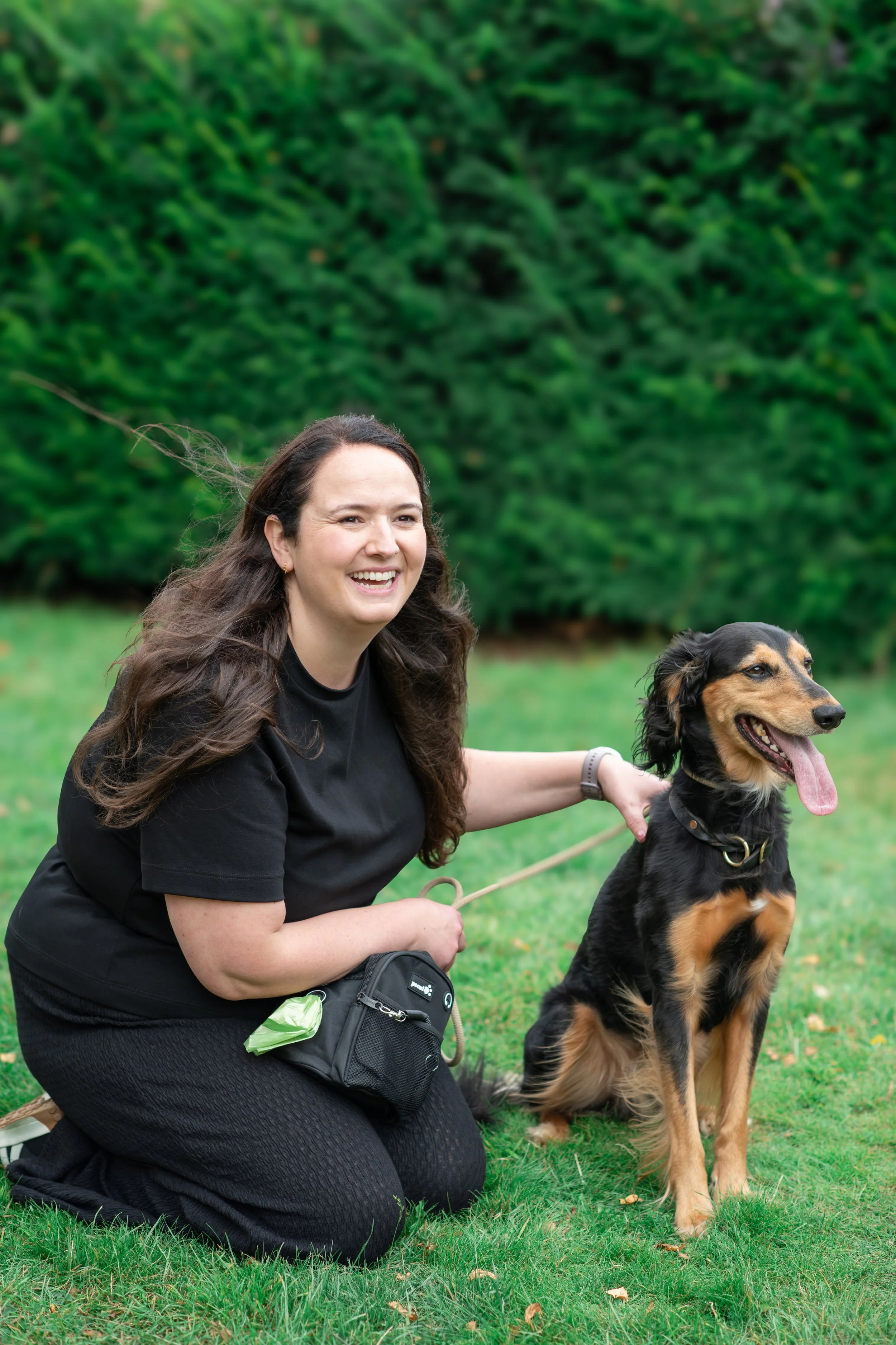 A woman kneeling on grass, smiling, with a black and brown dog sitting beside her. She is holding the dog's leash and outdoors with green bushes in the background.