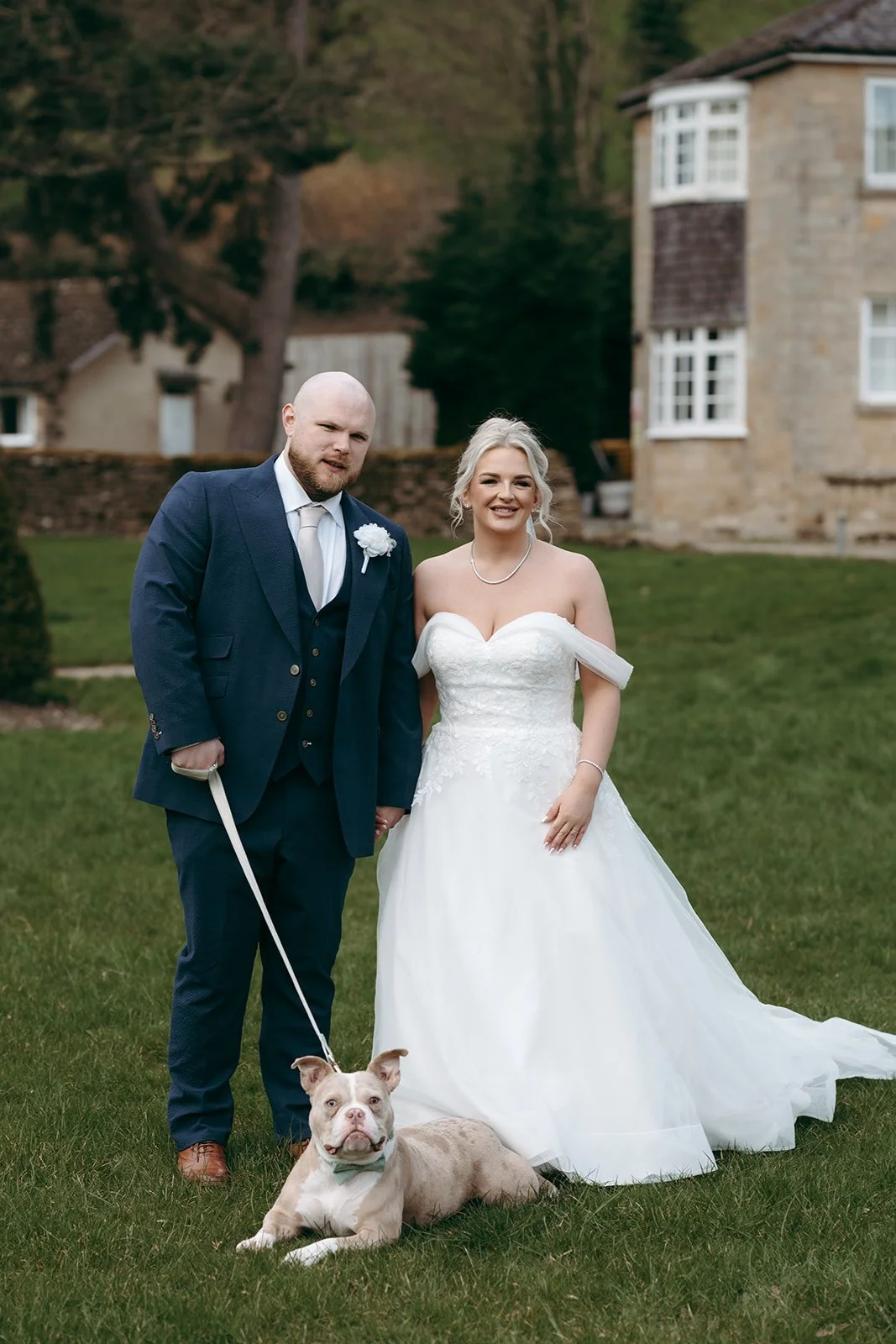 A newlywed couple standing on a green lawn with a dog lying down in front of them. The groom is wearing a dark blue suit with a boutonniere, and the bride is wearing a strapless white wedding gown with a veil. The background features a stone house an