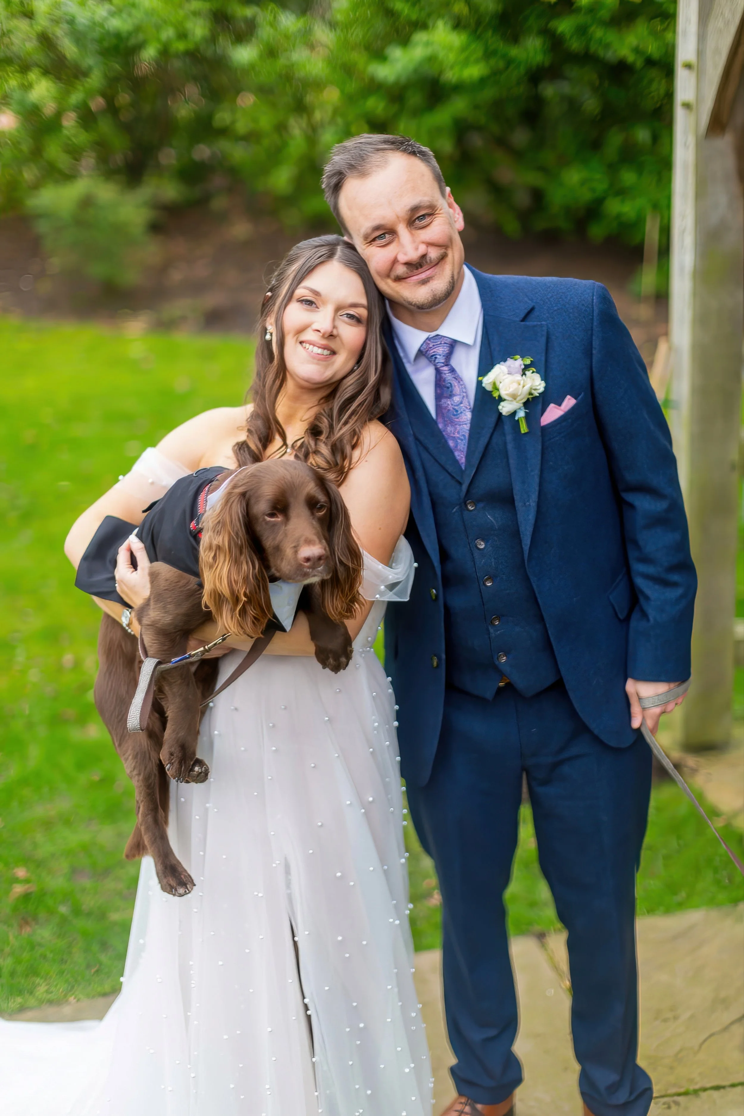 A couple dressed in wedding attire posing outdoors with a brown dog, greenery in the background, and smiling at the camera.