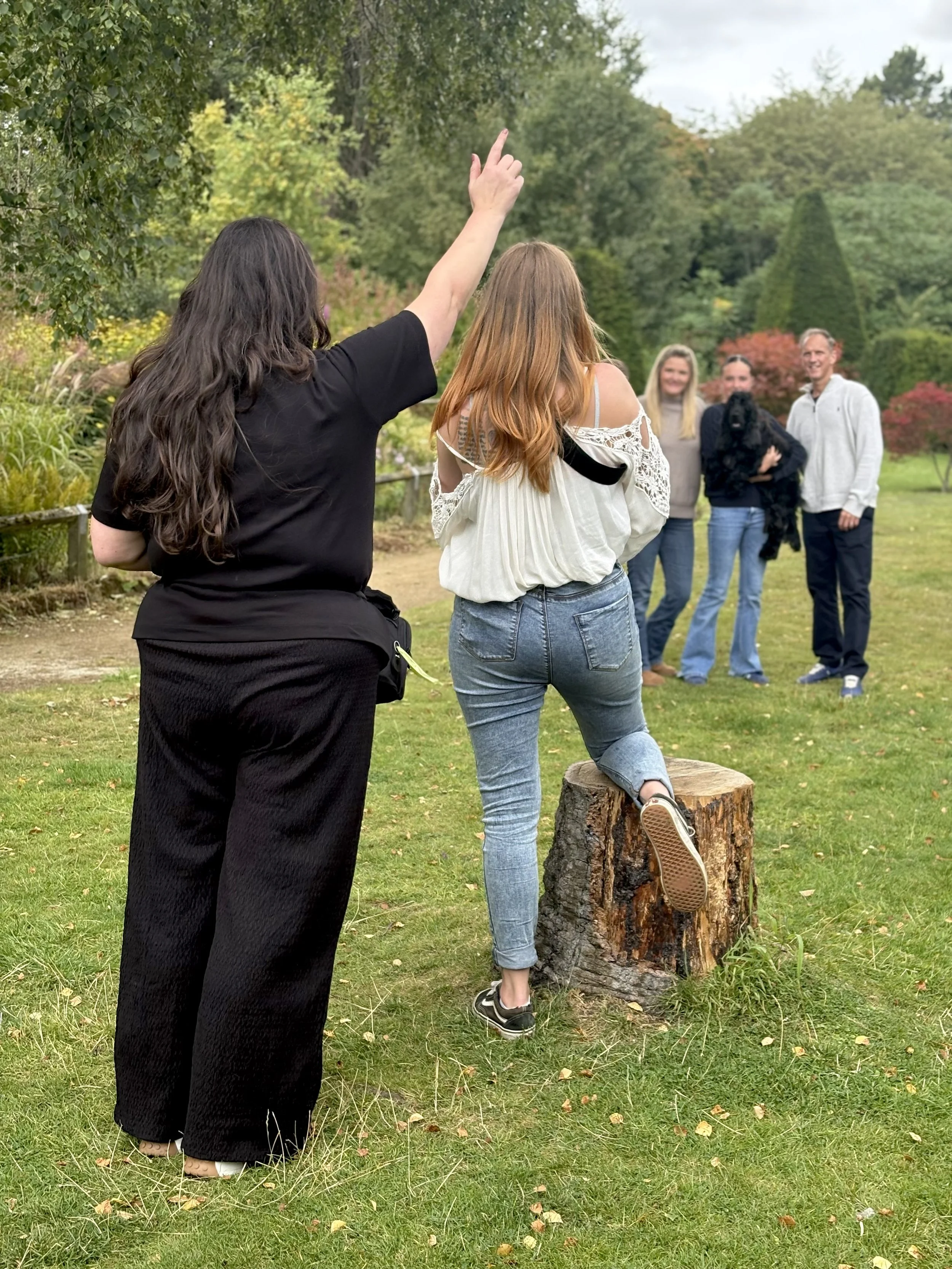 A group of five people and a dog in a park during autumn, with colorful trees in the background. One woman with long brown hair is standing with her back to the camera, raising her right arm, while another woman with red hair is balancing on a tree stump, facing away from the camera. Three people are standing in the background, smiling at each other, with a black dog being held by one of them.