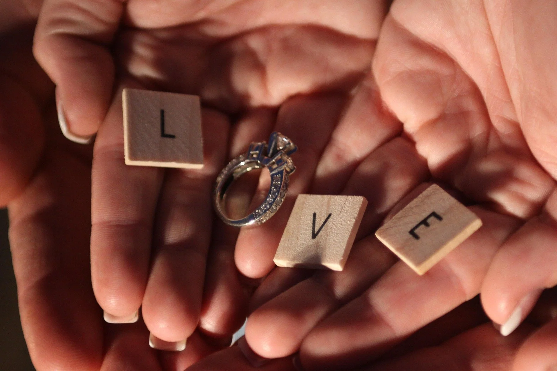A close-up of a hand holding Scrabble tiles spelling out 'LOVE' with a silver ring with embedded stones placed on the letter 'O' in the word.