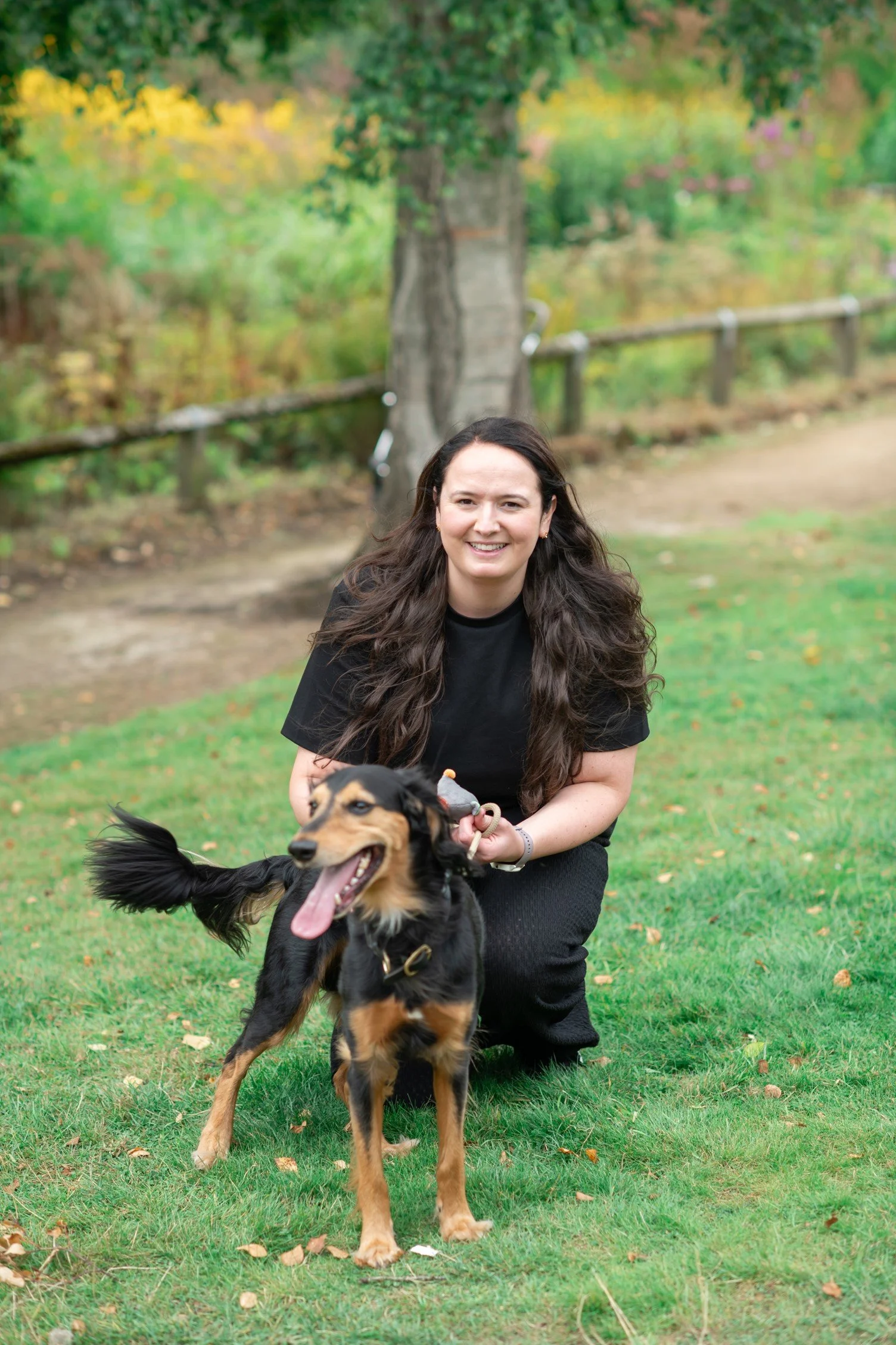 A woman with long brown hair smiling and kneeling on grass with her black and tan dog outdoors near a tree in a park.