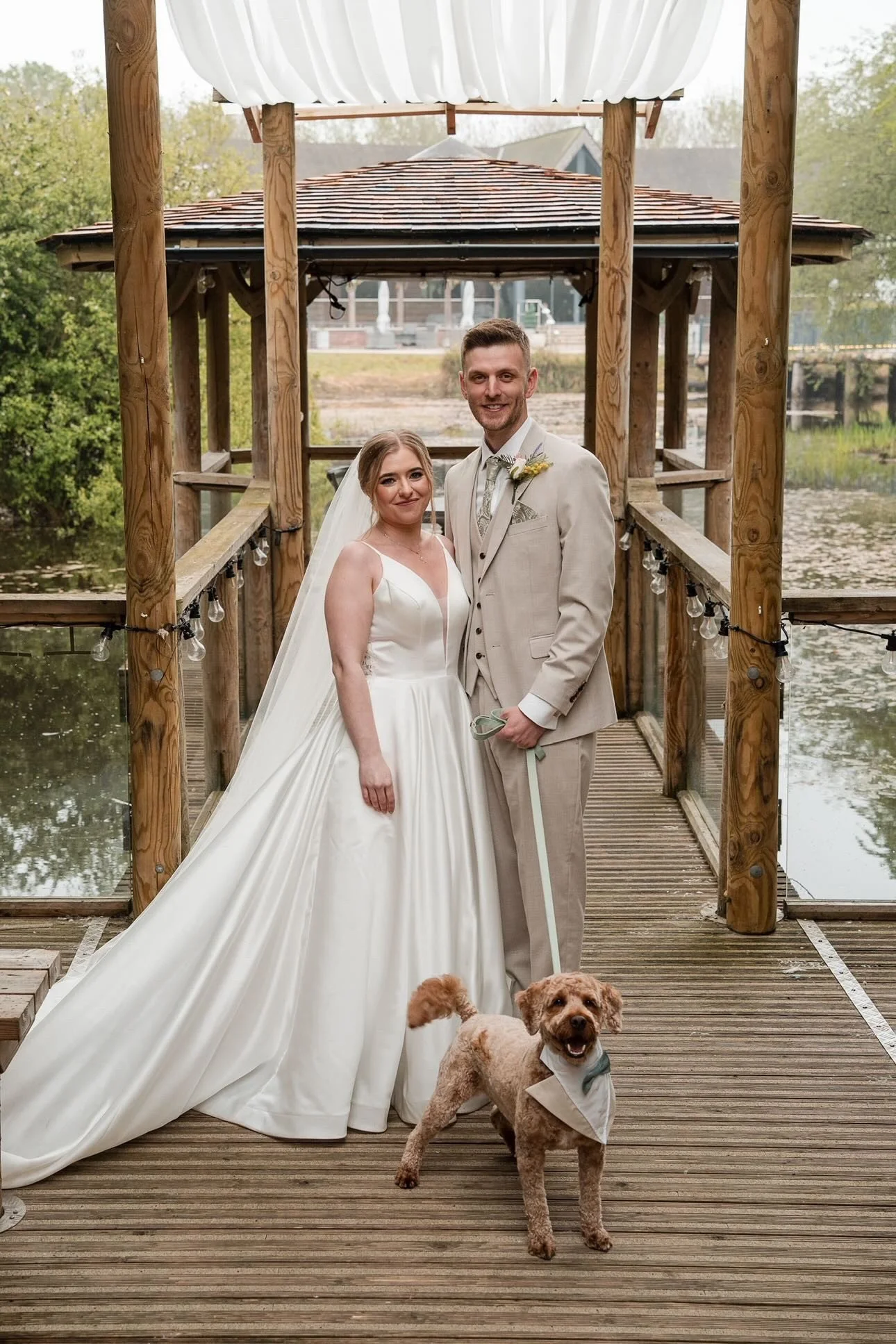 Bride and groom smiling with dog standing at their feet with a bow tie on