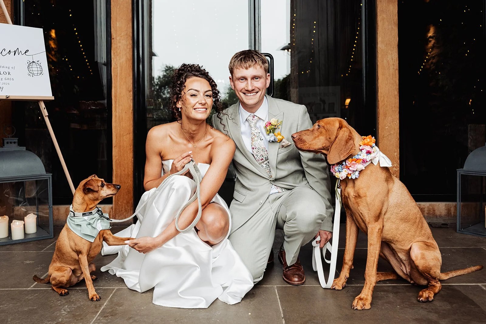 Two light brown dogs standing either side of the bride and groom. The larger dog wears a flower collar and the smaller dog wears a light green tuxedo and bow tie