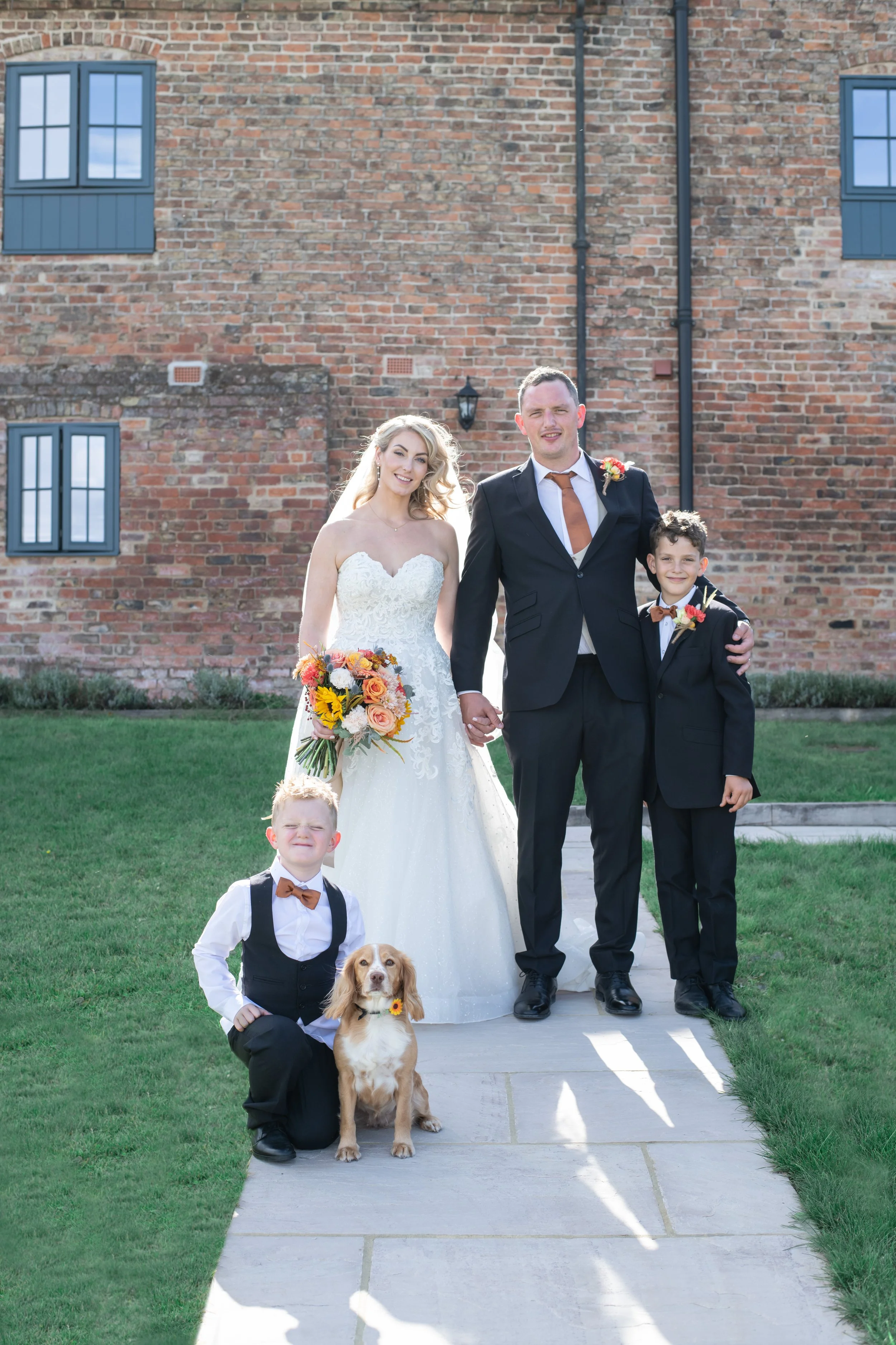 A wedding photo of a bride and groom with two young boys and a dog outside in front of a brick building. The bride is holding a bouquet of flowers, the groom is wearing a suit, and the boys are dressed in formal suits with bow ties.