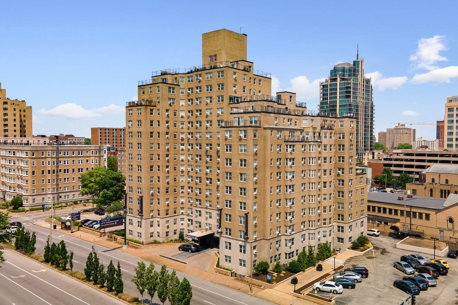 Tall beige apartment building with multiple balconies in an urban cityscape, surrounded by other buildings, trees, a parking lot, and a street with cars under a partly cloudy sky.