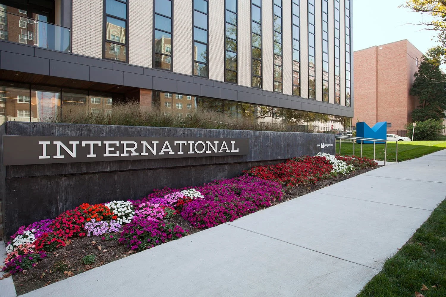 Exterior view of a modern office building with large windows, a landscaped flower bed with pink, white, and red flowers, and a sign that reads "INTERNATIONAL." There is a sidewalk adjacent to the flower bed and a blue logo in the background.