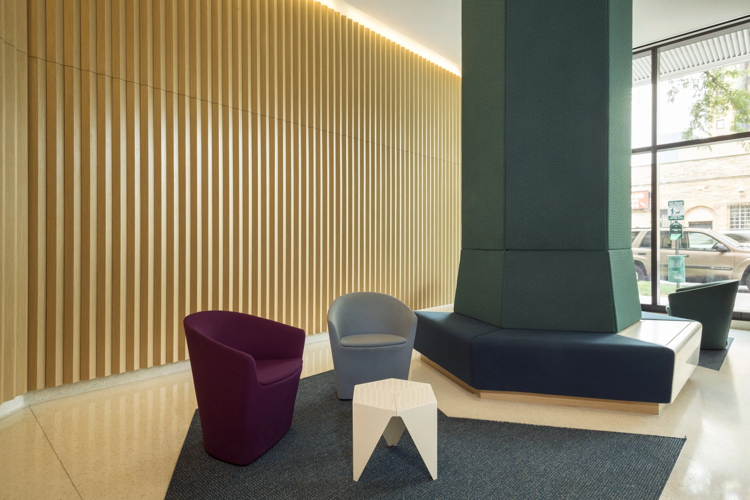 Modern interior with colorful chairs, a white small side table, wooden slatted wall, large green structure, and glass windows showing cars outside.