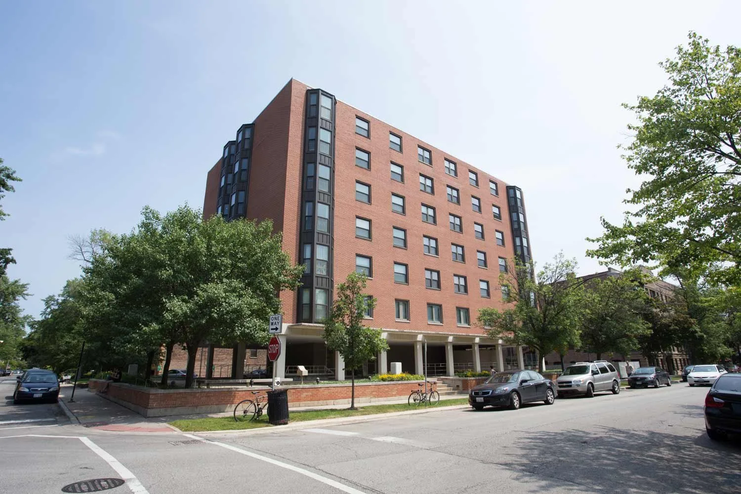 A multi-story brick apartment building with black window frames, surrounded by existing green trees and parked cars on the street in front.