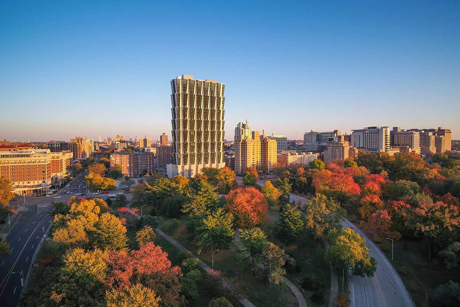 Cityscape with modern high-rise building, surrounded by colorful autumn trees and lower buildings under a clear blue sky.