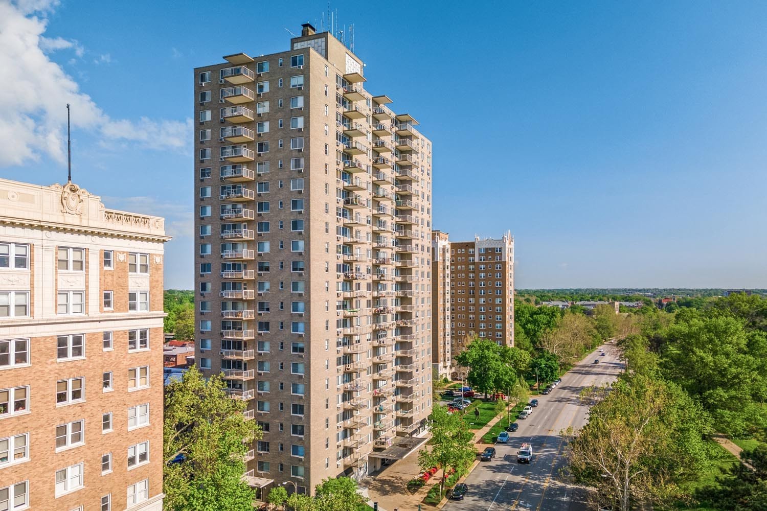 A cityscape with tall residential apartment buildings, a tree-lined street, and a blue sky with some clouds.