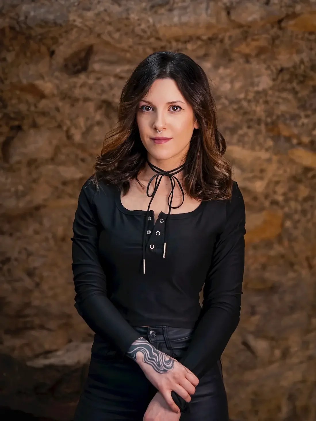 Une femme souriante aux cheveux bruns, portant un haut noir avec un collier à cordon et un tatouage sur la main, devant un mur en pierre.