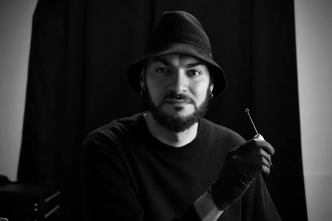 Un homme portant un chapeau noir et des boucles d'oreilles, tenant une aiguille à tatouage, regardant l'objectif, dans un environnement sombre.