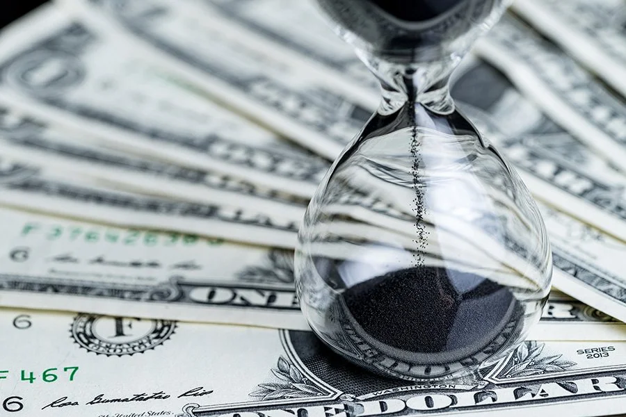 An hourglass with black sand on top of dollar bills, symbolizing the concept of time and money.