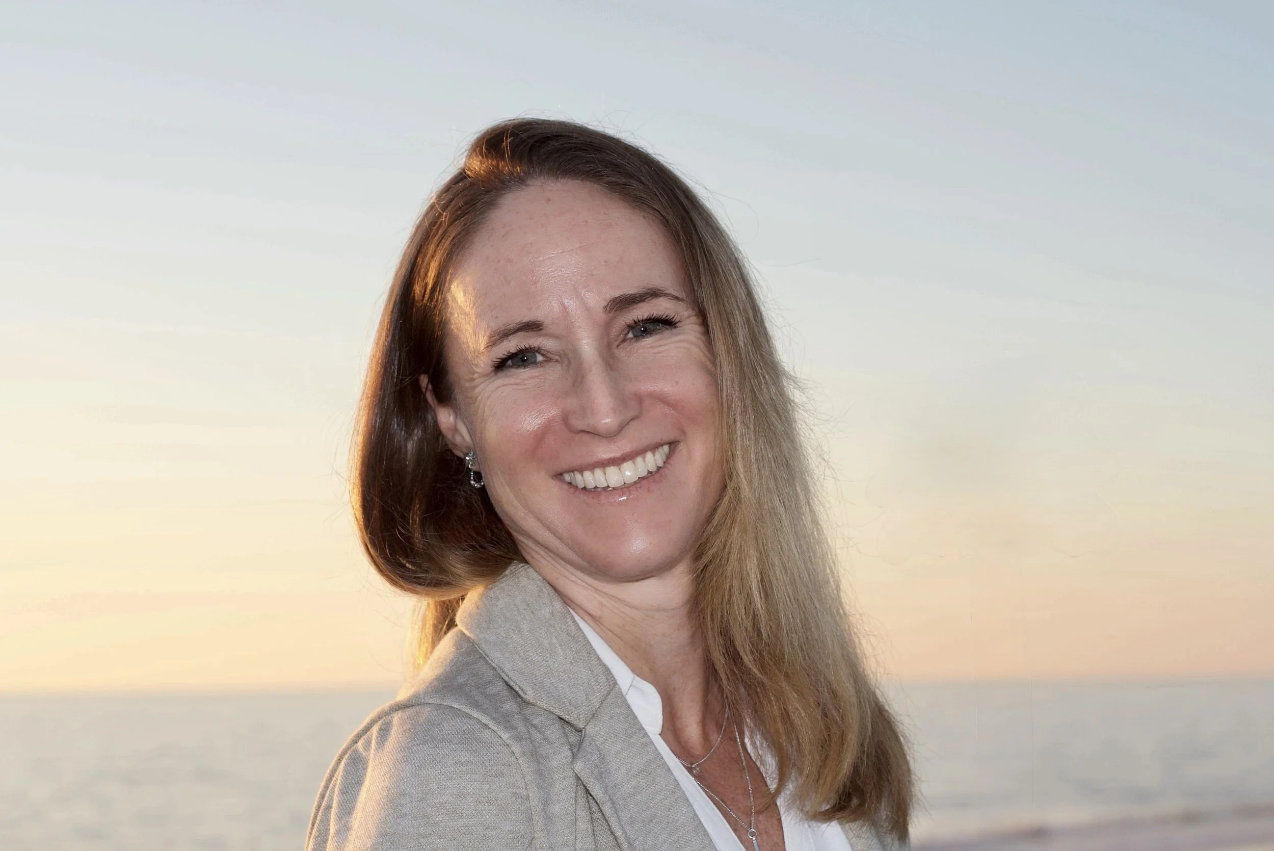 A woman smiling outdoors at sunset or sunrise with the ocean in the background.