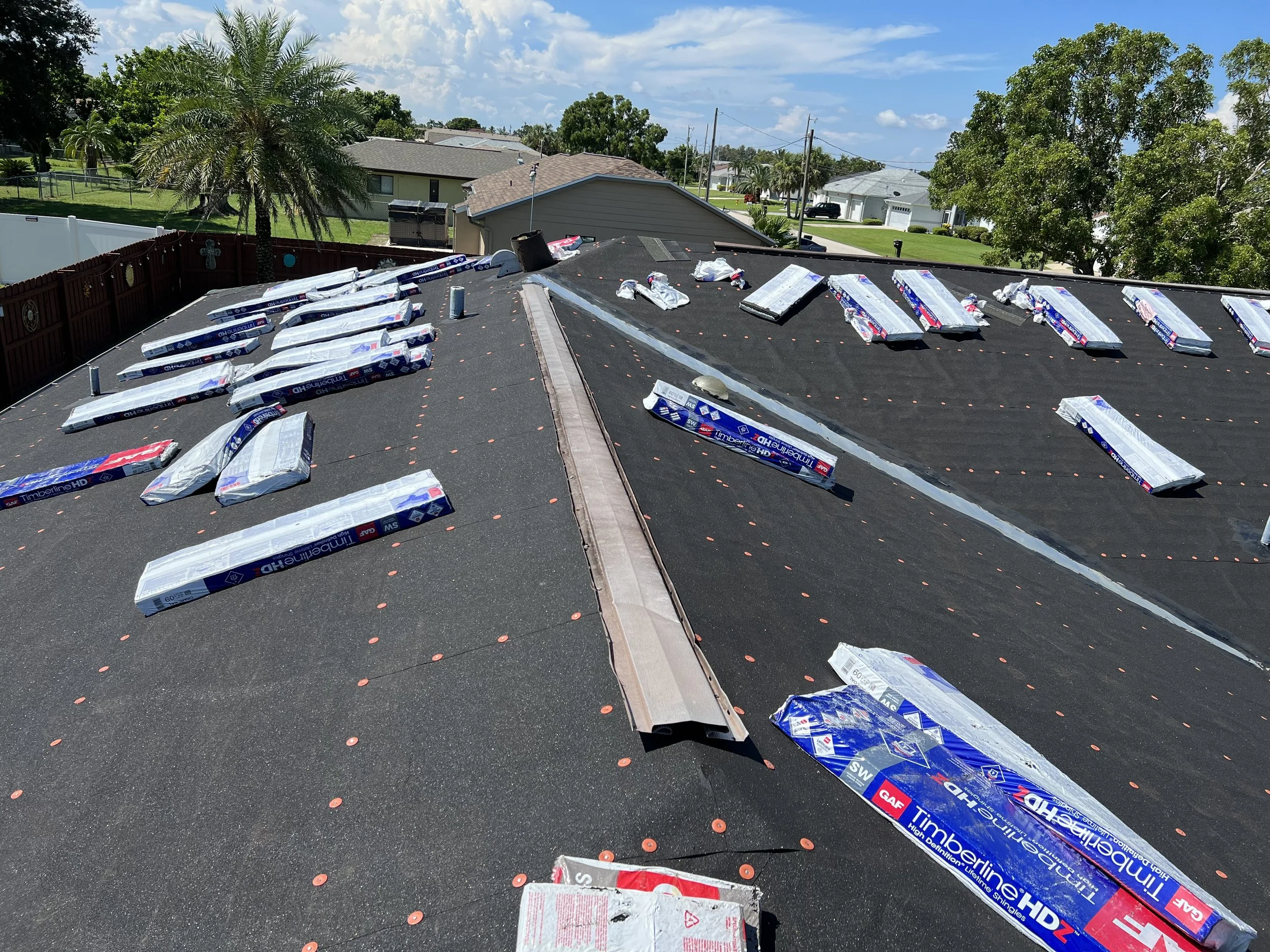 Roof with shingles and supplies scattered on top, residential neighborhood background under blue sky.