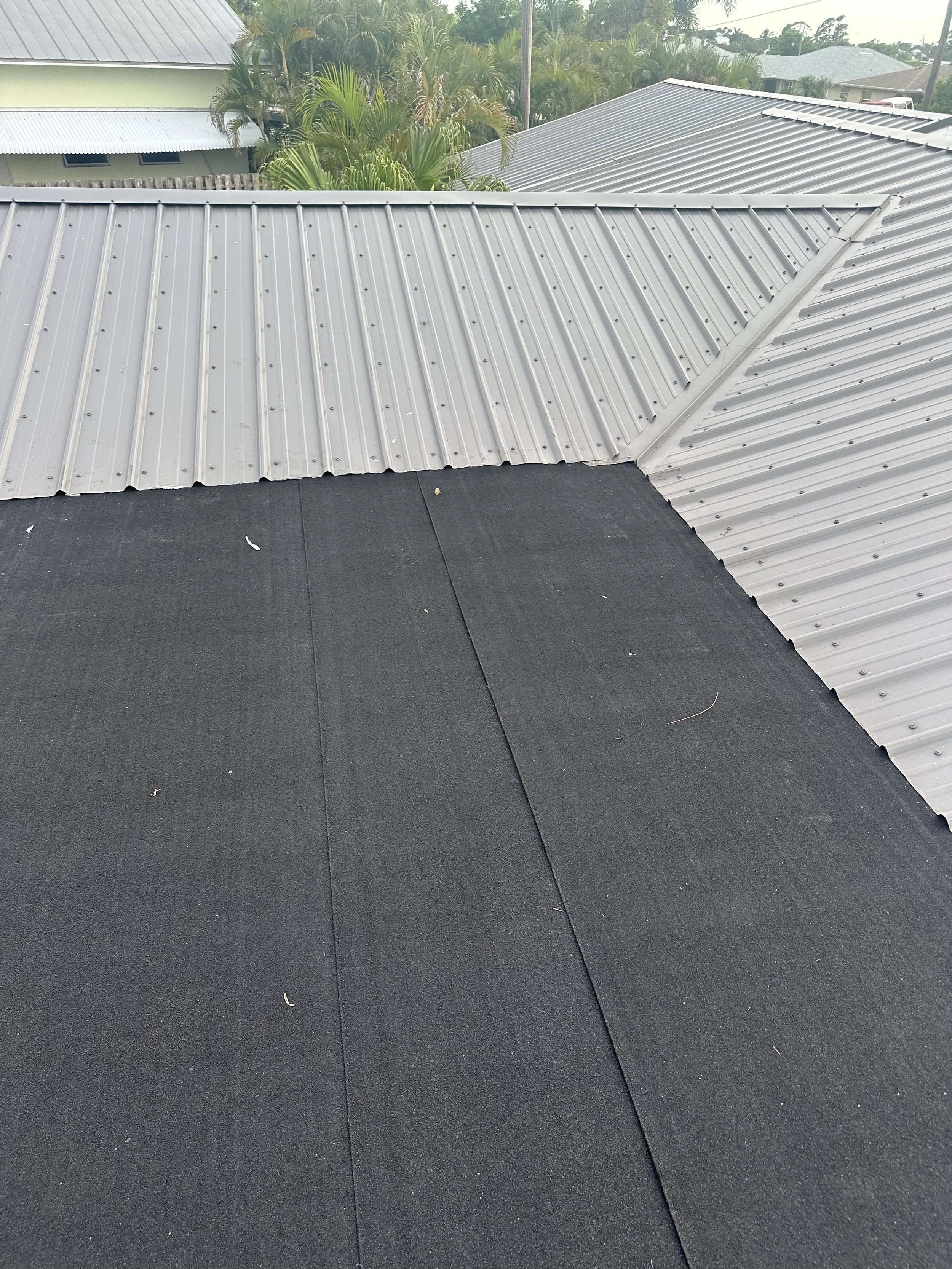 Top view of a roof with black asphalt shingles and silver metal panels, surrounded by green palm trees and neighboring houses under a cloudy sky.