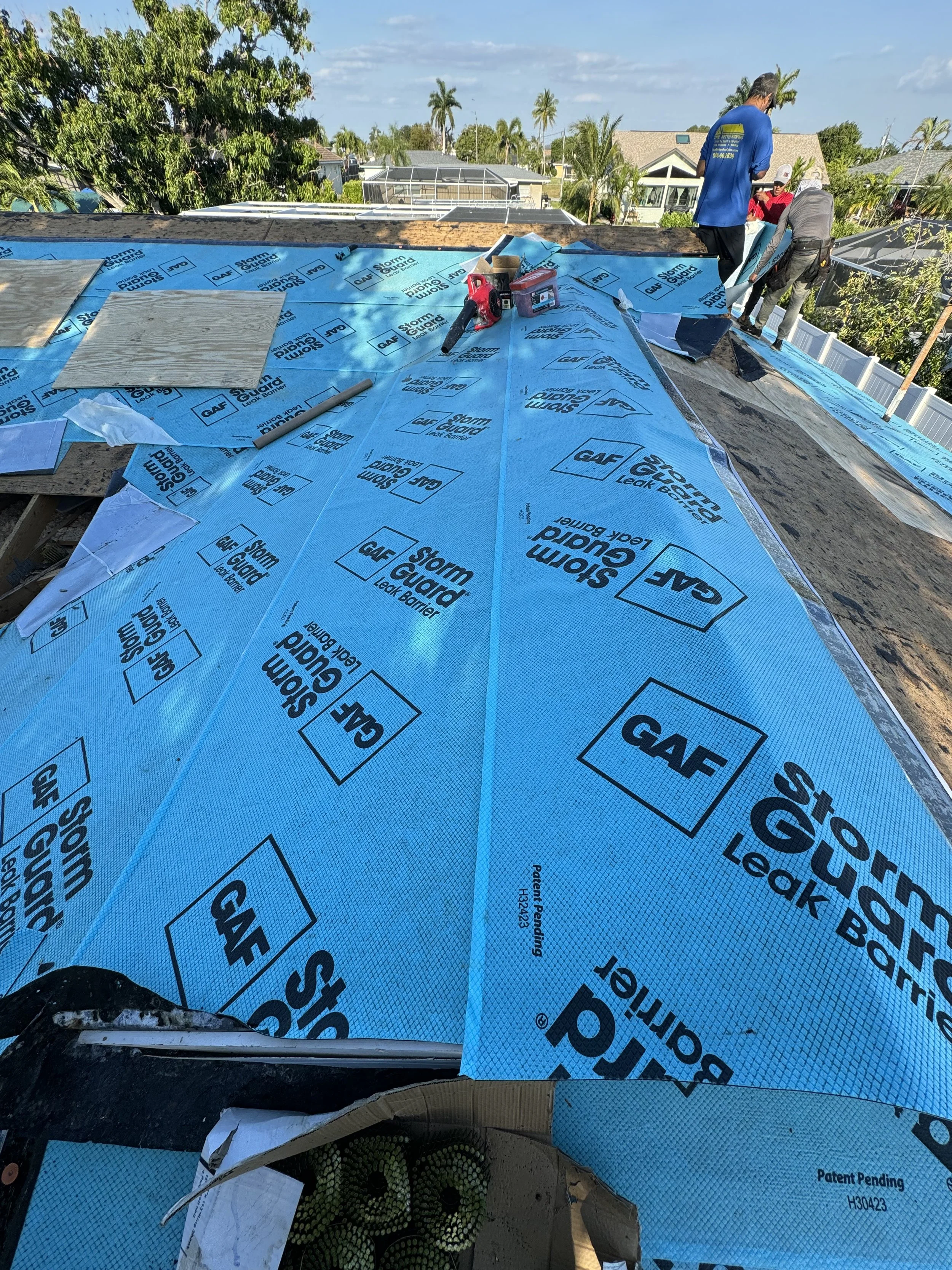 Roof under construction covered with blue waterproof Storm Guard Leak Barrier material, with workers installing roofing components on a residential house in a suburban neighborhood with palm trees and other houses in the background.