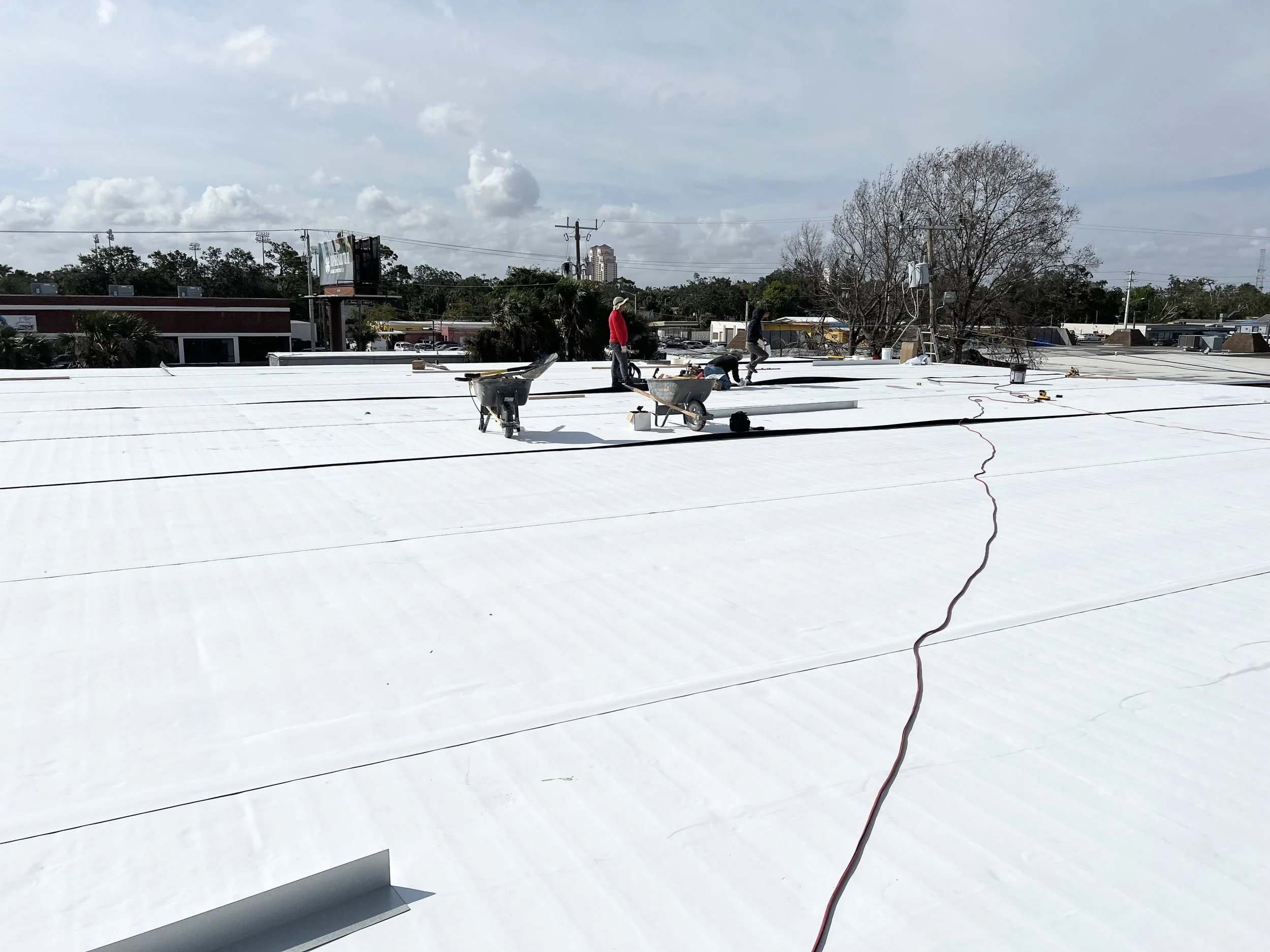 Workers installing a white reflective roof on a commercial building under partly cloudy sky.