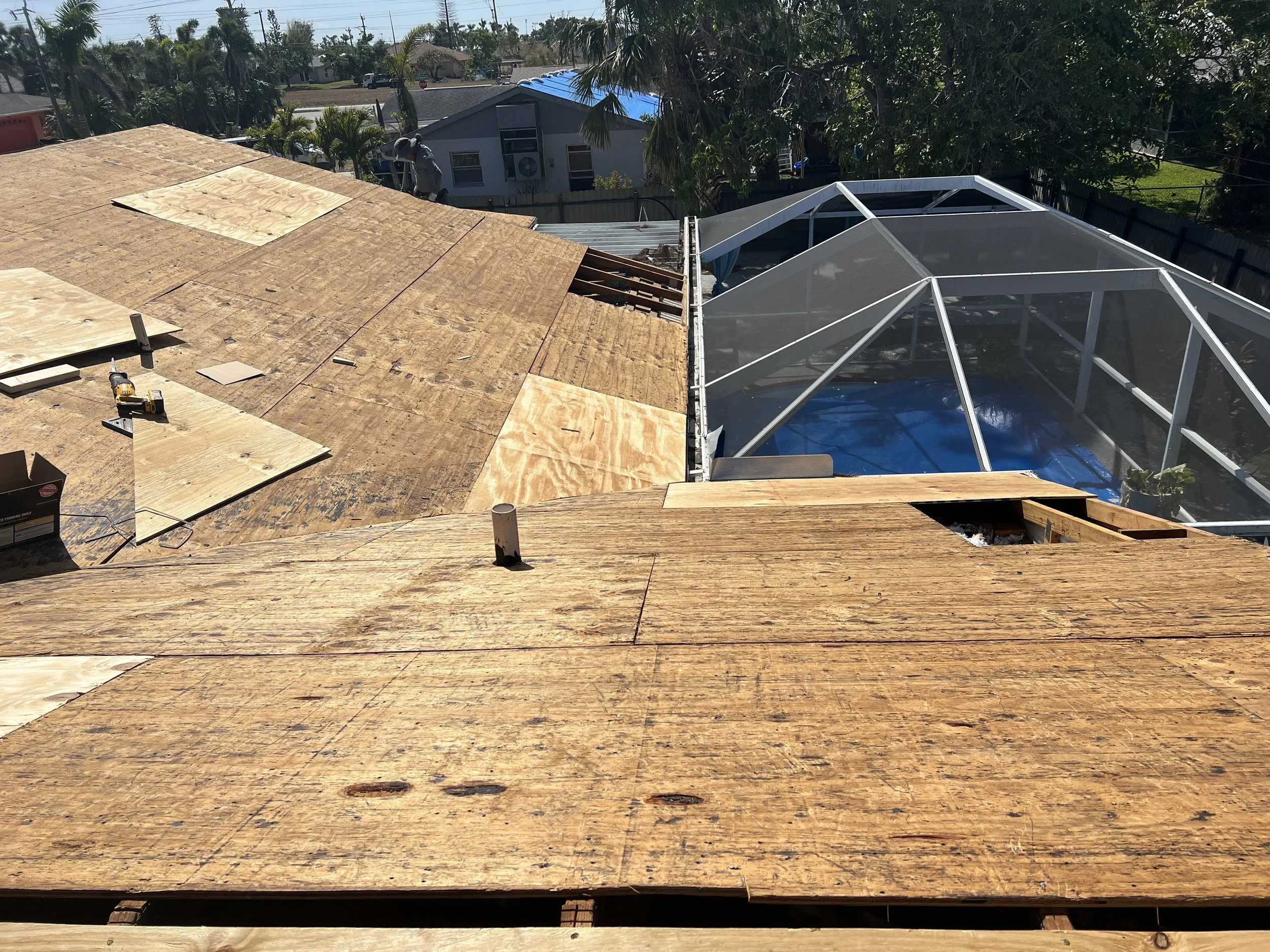Building roof under construction with wooden boards and a partially installed glass or plastic enclosure, blue tarp on ground inside the enclosure, tools and materials scattered on the roof.