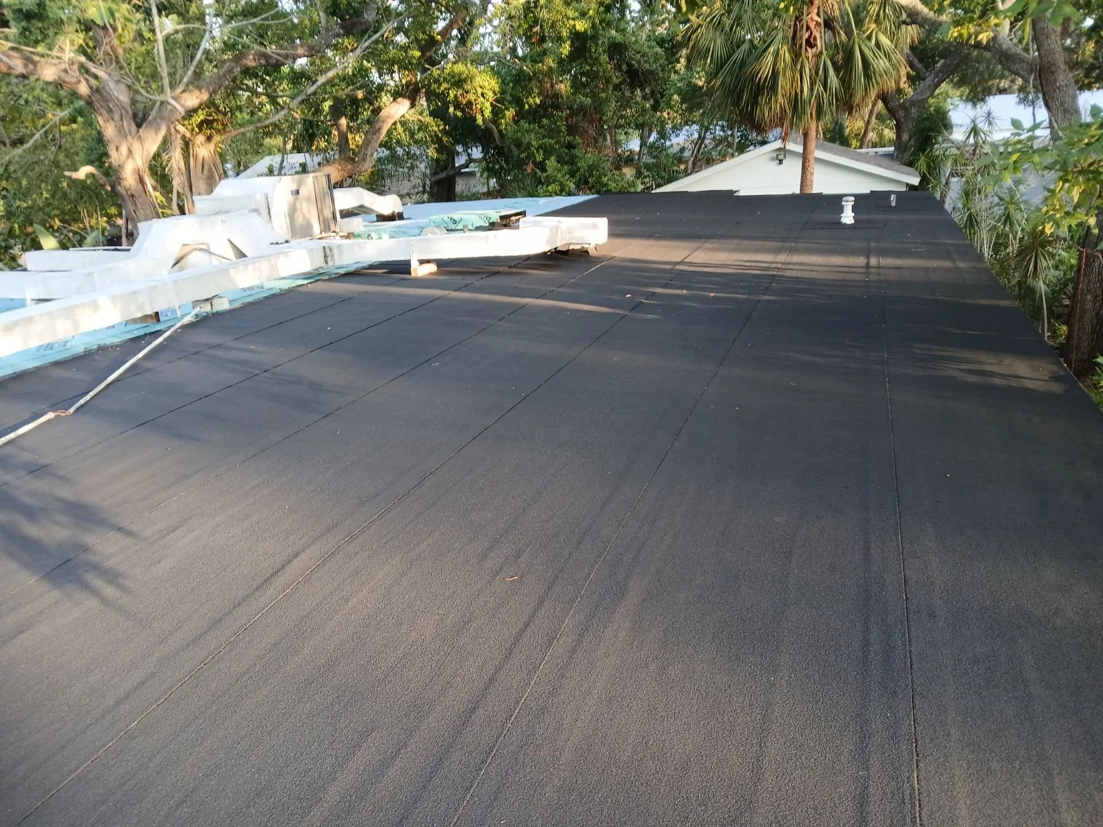 View of a roof with black roofing material and some construction materials on top, trees in the background, and neighboring houses.