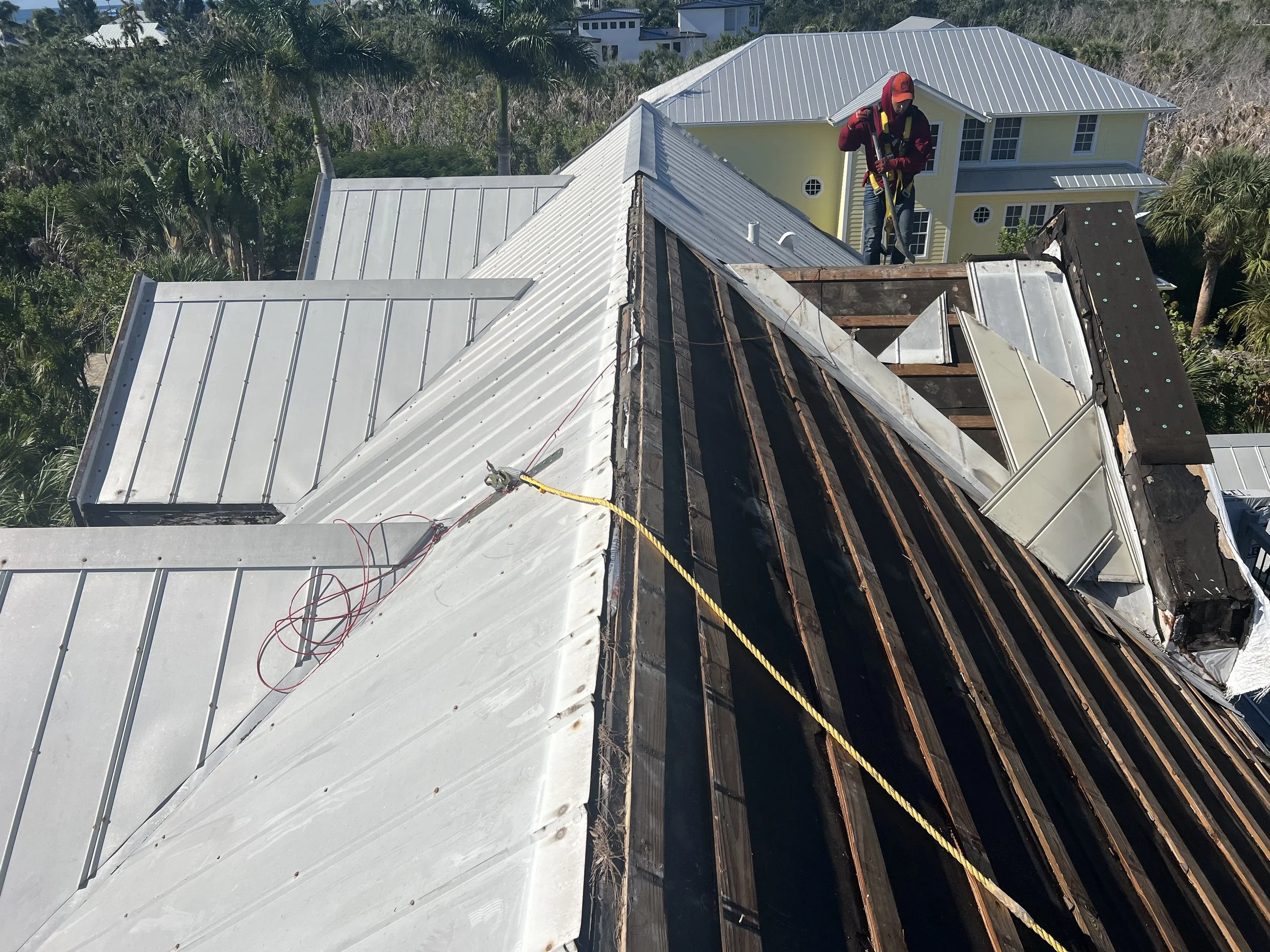 A worker stands on a roof under repair, wearing safety gear and examining the roofing structure. The roof has sections with roofing panels and exposed wooden beams.