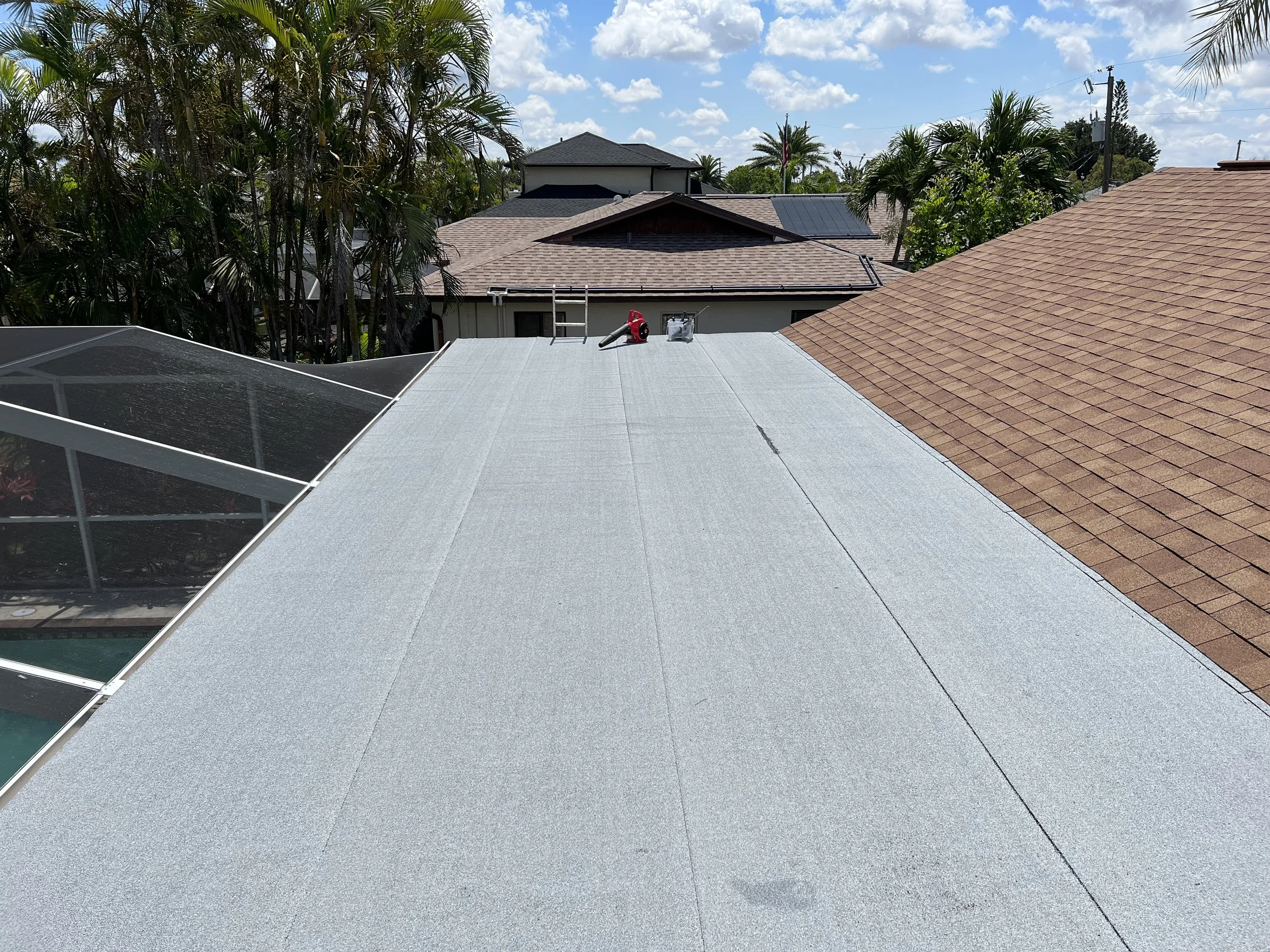 View from the roof showing multiple neighboring houses with shingle and tile roofs, a screened pool area on the left, trees, and a partly cloudy sky.