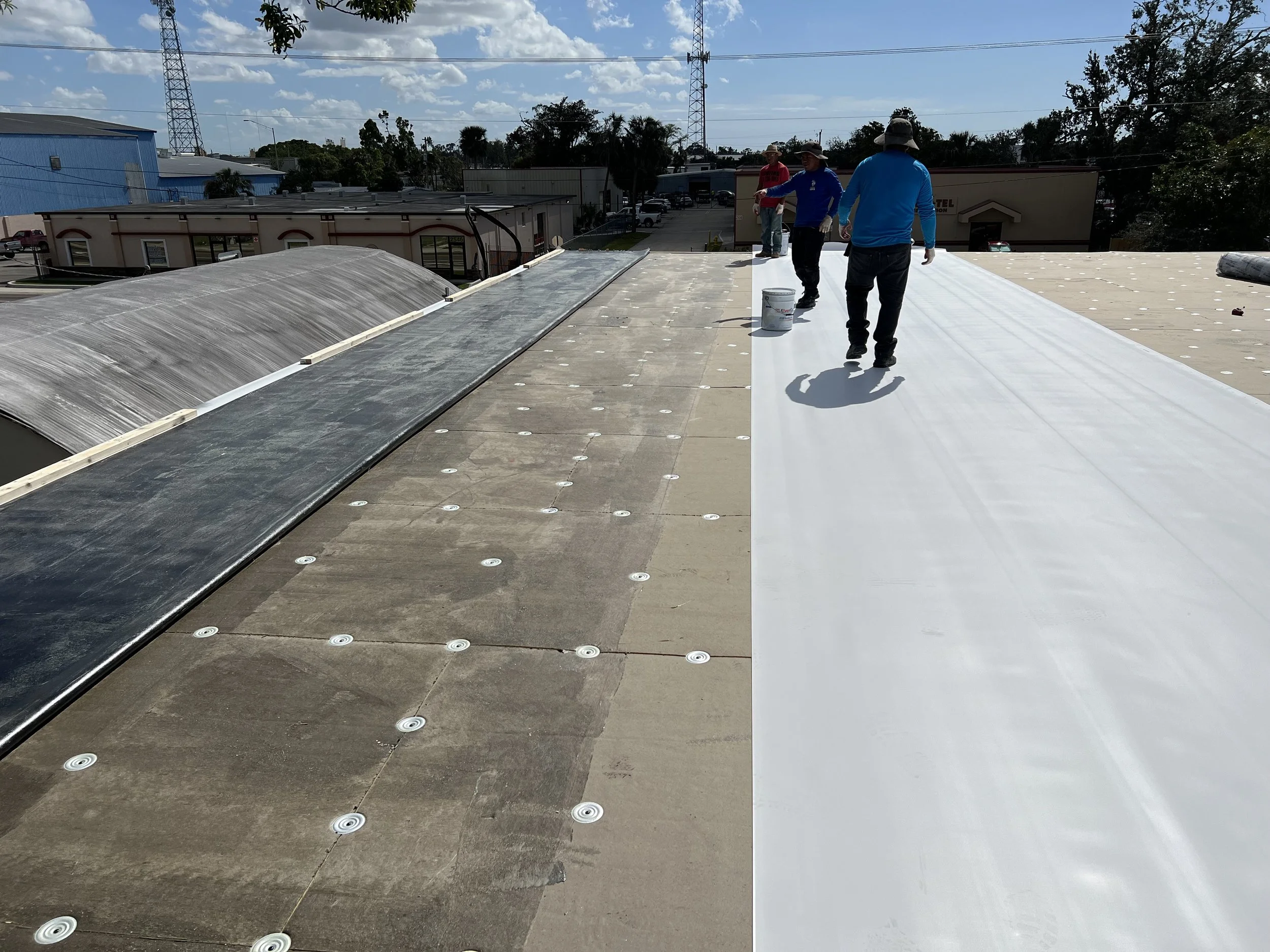 Construction workers installing a new white roofing material on a commercial building's roof. The sky is partly cloudy.