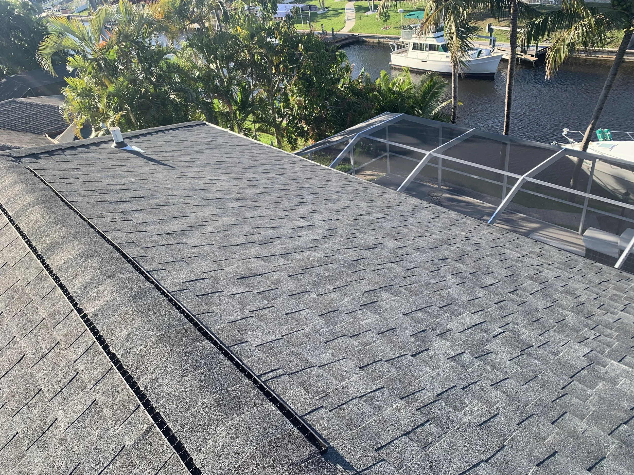 View of a gray shingled rooftop overlooking a backyard with palm trees, a canal, and boats, including a covered area with a screened porch.