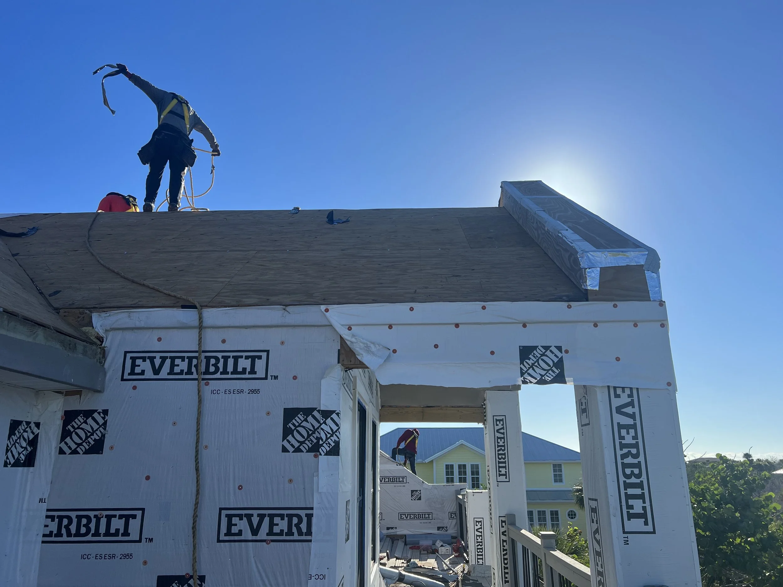 Two workers on a house under construction, one on the roof and another in the background, with blue sky and sunlight.