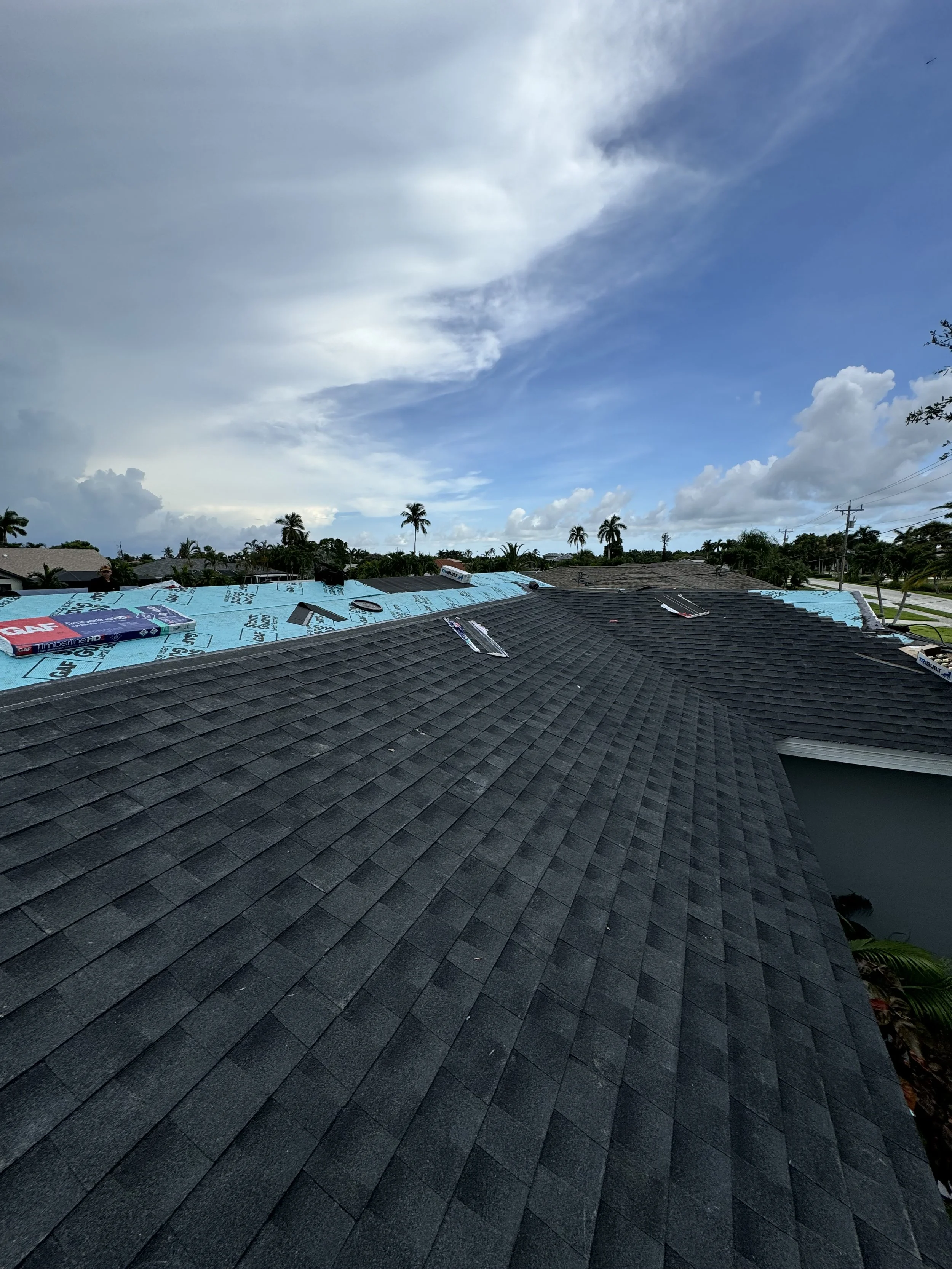 Residential roof with shingles, partially under repair with blue roofing underlayment visible, and tools and materials on top. Palm trees and a partly cloudy sky in the background.