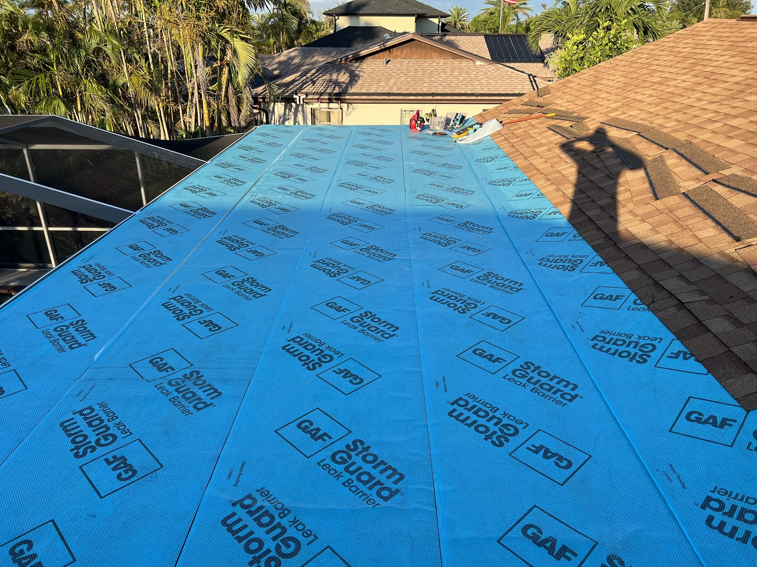 A rooftop under construction with blue storm guard leak barrier material. The roof has brown shingles on one side and a new section being covered with storm guard. There are tools and materials on top of the roof. The background shows neighboring hou