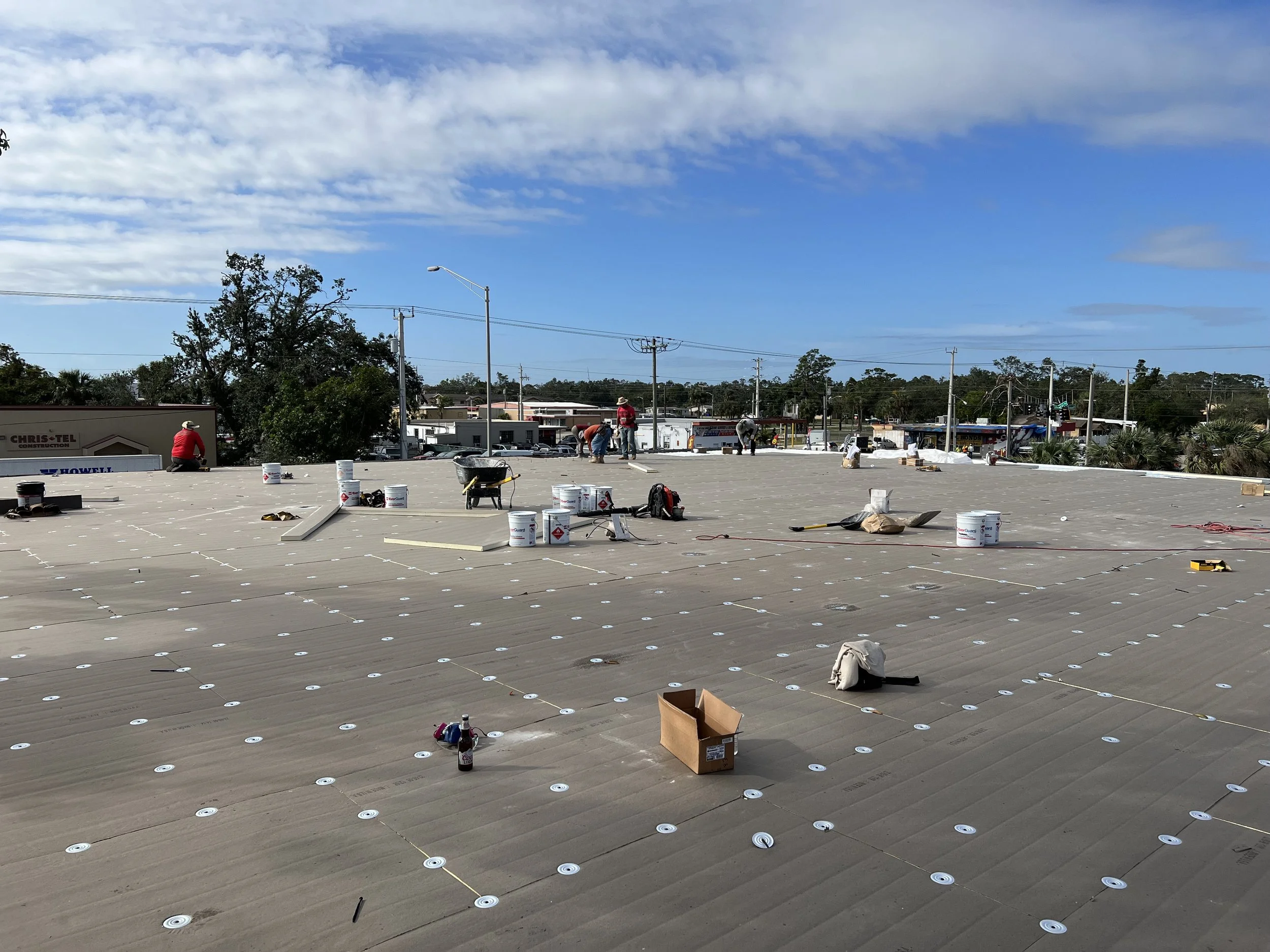 Workers installing a new roofing system on a commercial building. The flat roof is covered with large, gray roofing panels, and various tools and buckets are scattered across the surface under a partly cloudy sky.