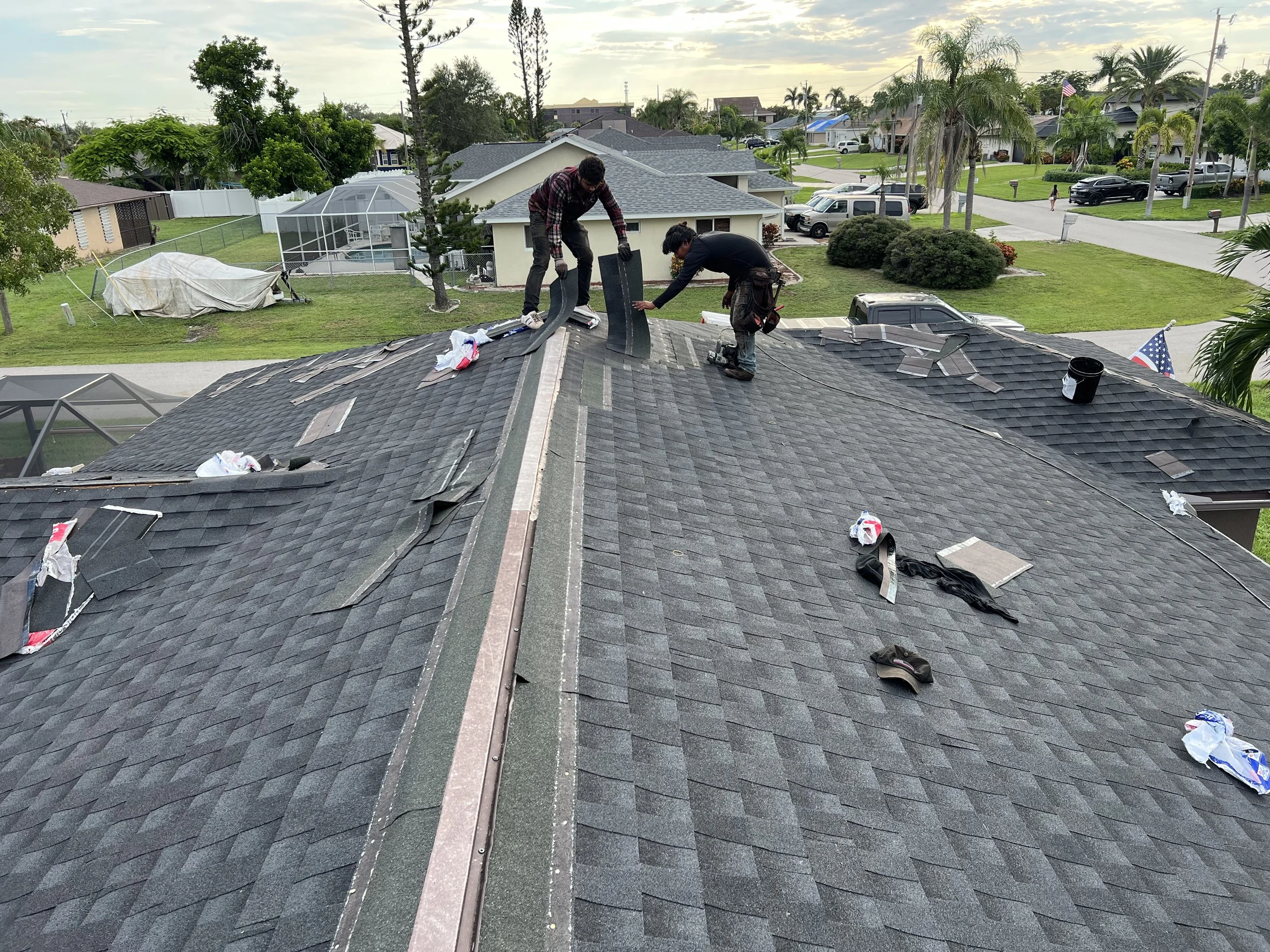Two workers on a house roof installing or repairing shingles, surrounded by roofing tools and materials, with a suburban neighborhood in the background.