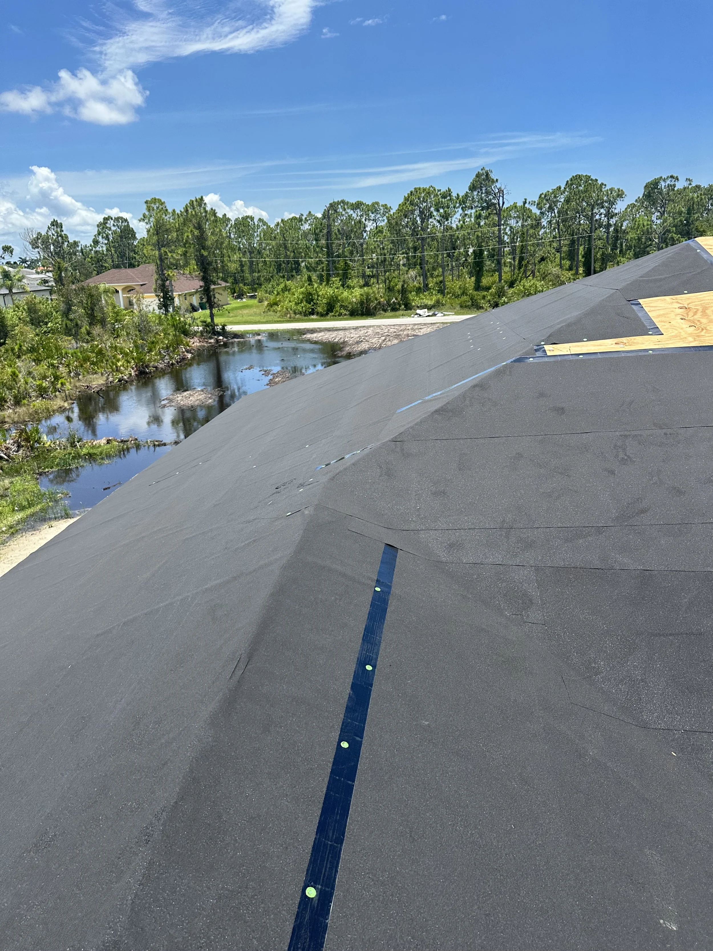View of a roof under construction with black roofing material and blue sealant strip, in a residential neighborhood with trees, water, and blue sky in the background.