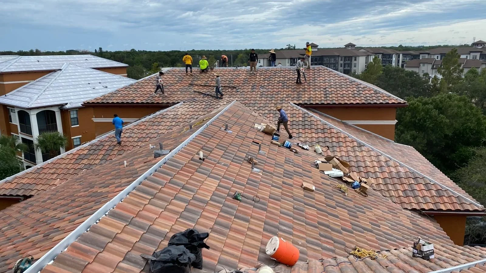 Roofers working on a large multi-section tiled roof, scattered with tools and materials, with trees and apartment buildings in the background.