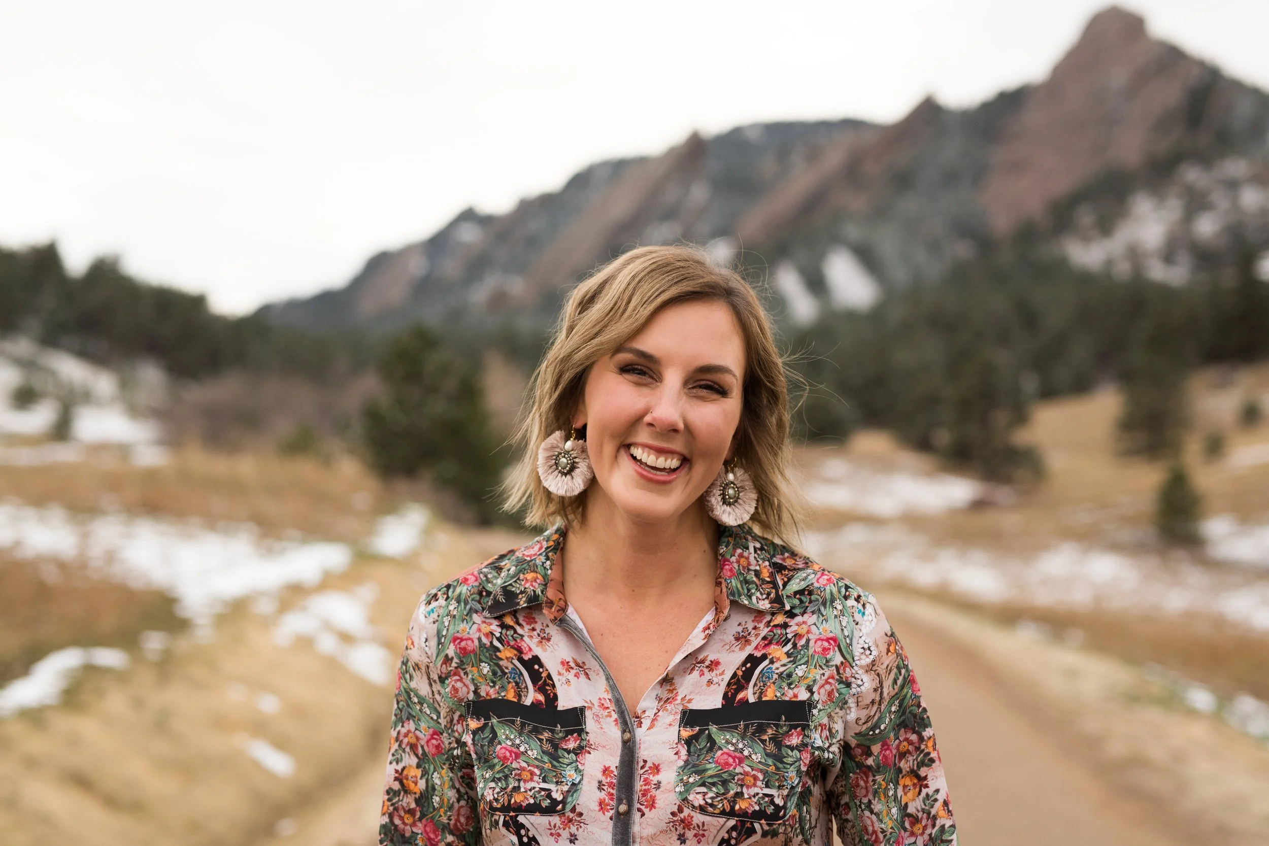 A woman with short blonde hair smiling outdoors in a mountainous area with patches of snow and trees, wearing large earrings and a floral jacket.
