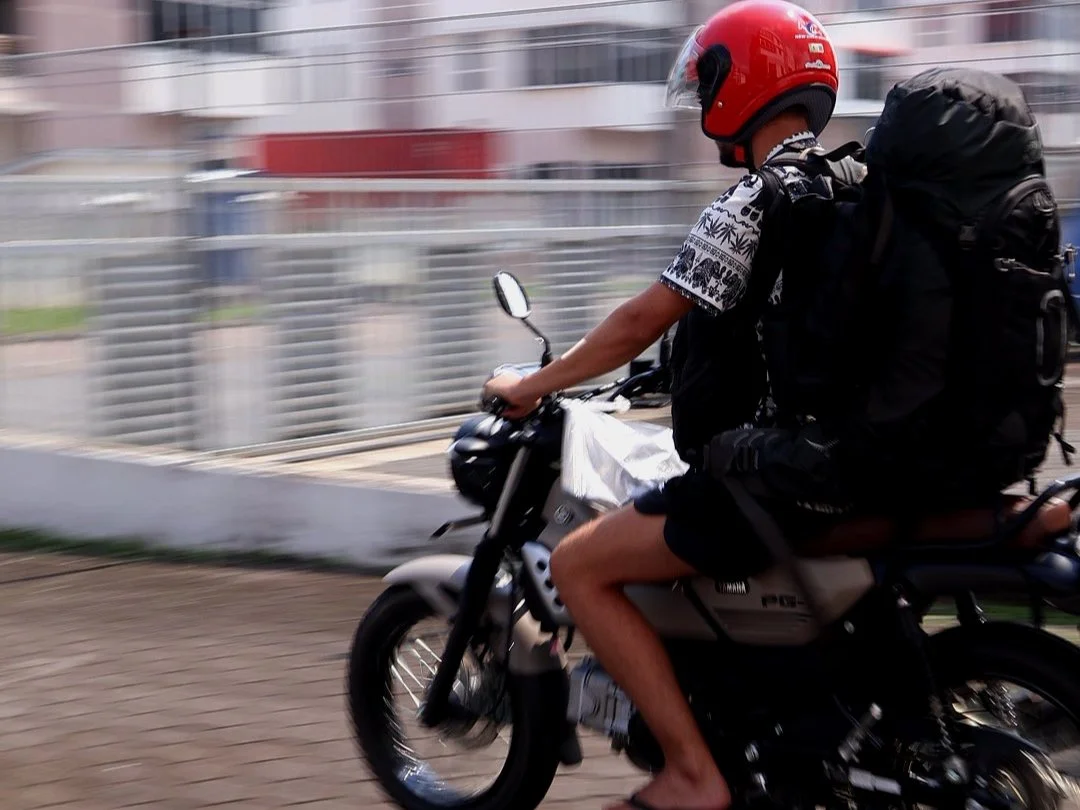 Young person riding a motorcycle with a large backpack, wearing a red helmet, t-shirt, shorts, and sandals, in an urban environment.