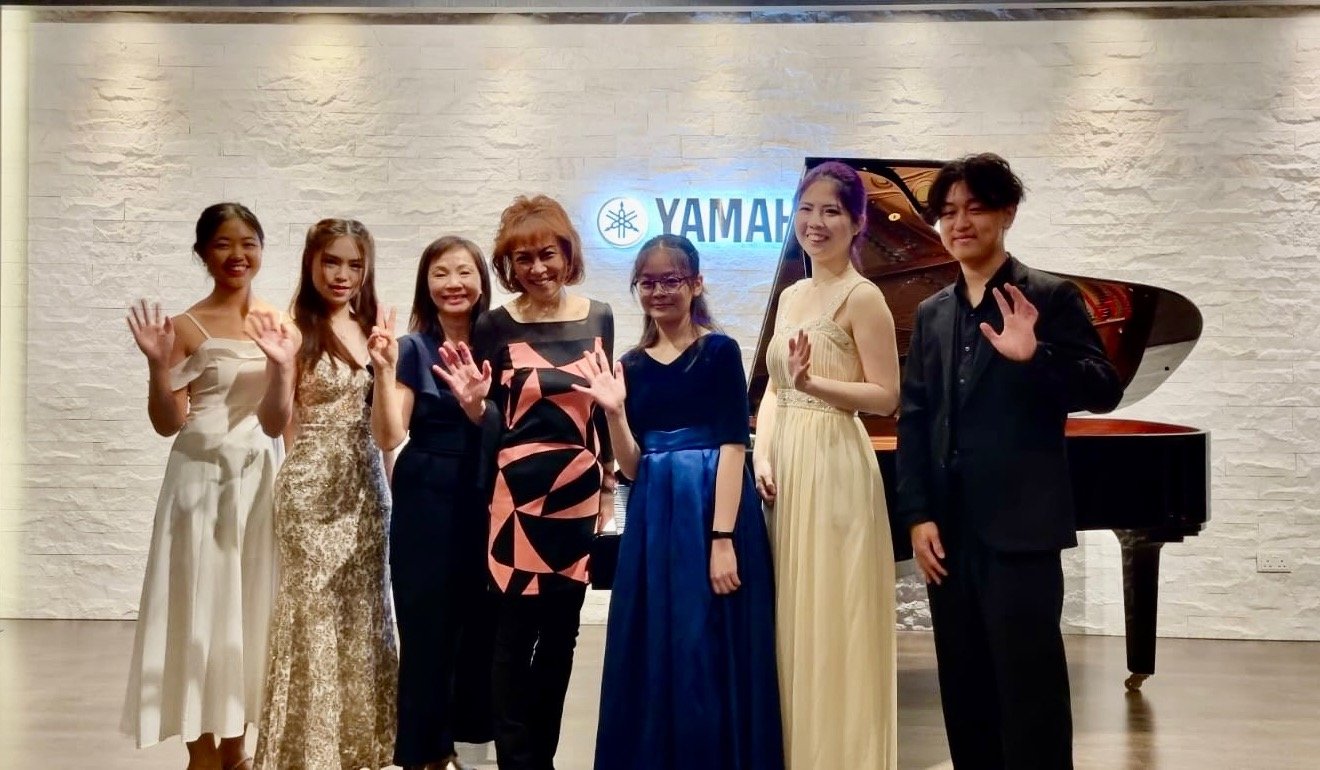 Group of seven people posing in front of a grand piano, with a Yamaha logo on the wall behind them, in an indoor venue with white brick walls.