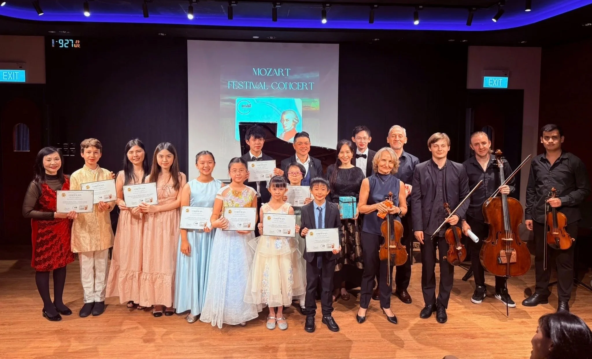 A group of children and adults on stage at a concert hall, holding certificates and musical instruments, after a concert performance with a screen behind them displaying the text 'Mozart Festival Concert'.
