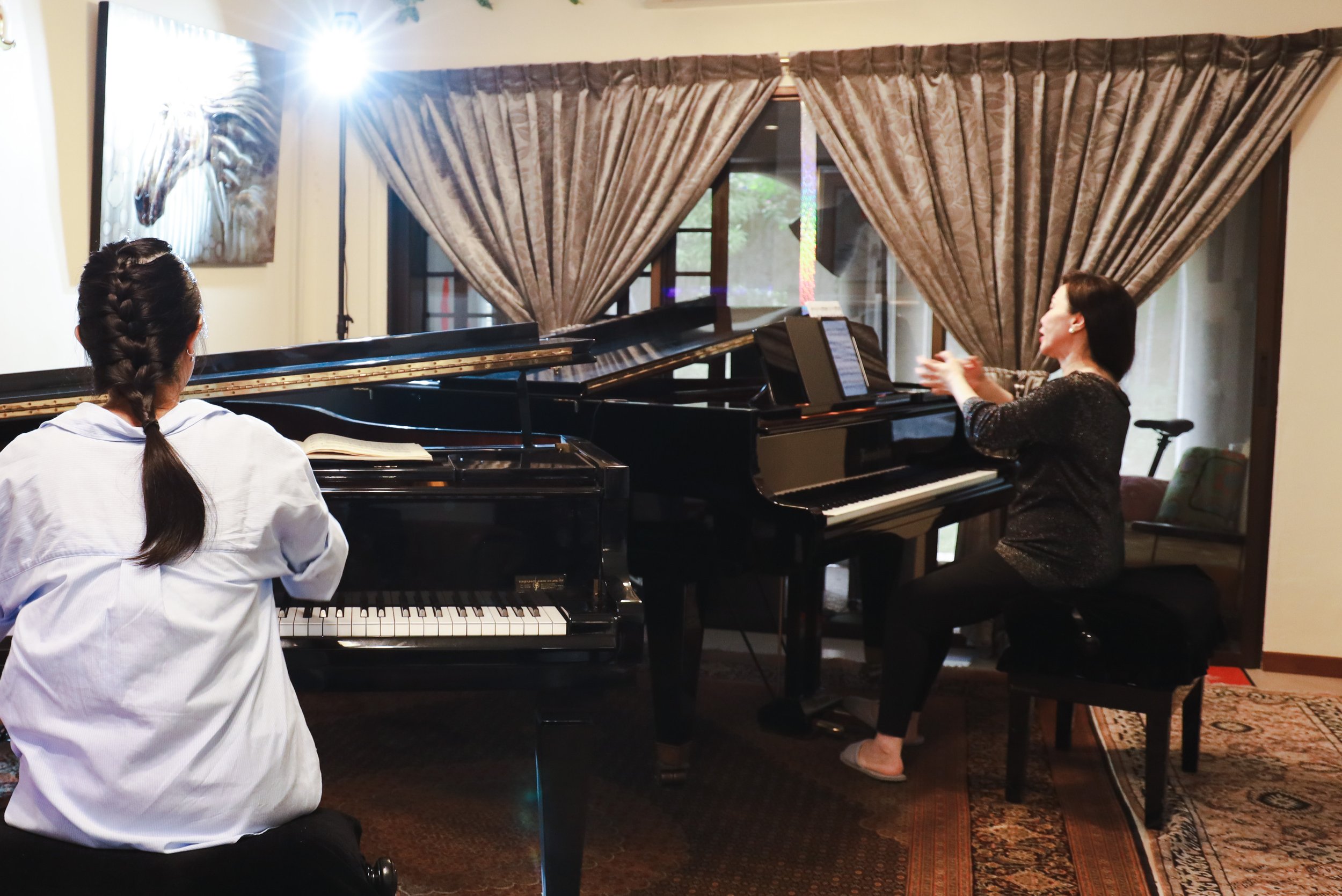 Two women are in a room with grand pianos, one playing the piano and the other observing, with a large window, curtains, artwork, and a patterned rug in the background.