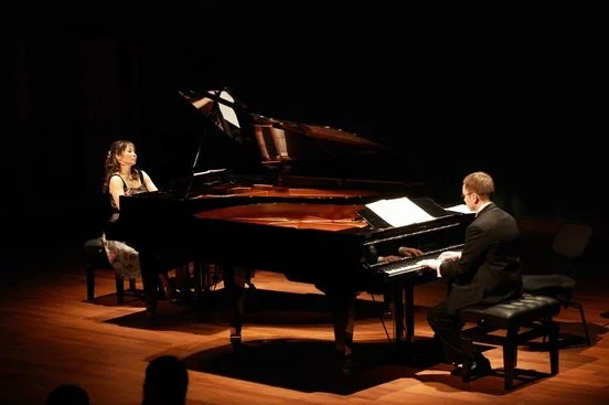 Two pianists, a woman and a man, performing a duet on a grand piano on a stage with a dark background.
