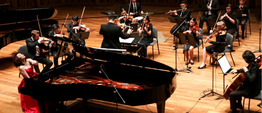 Orchestra performing on stage with a grand piano, conductor, and musicians playing string instruments, including violins and cello.