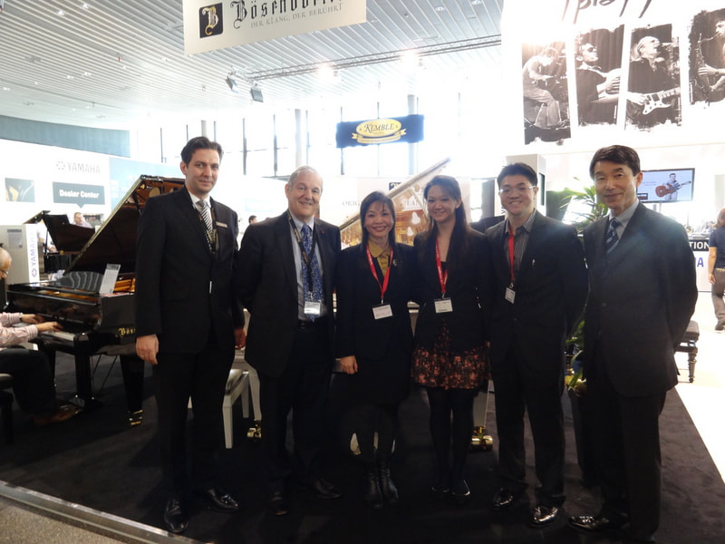 Group of five people standing in front of a piano and a large black and white poster at an indoor event or exhibition.
