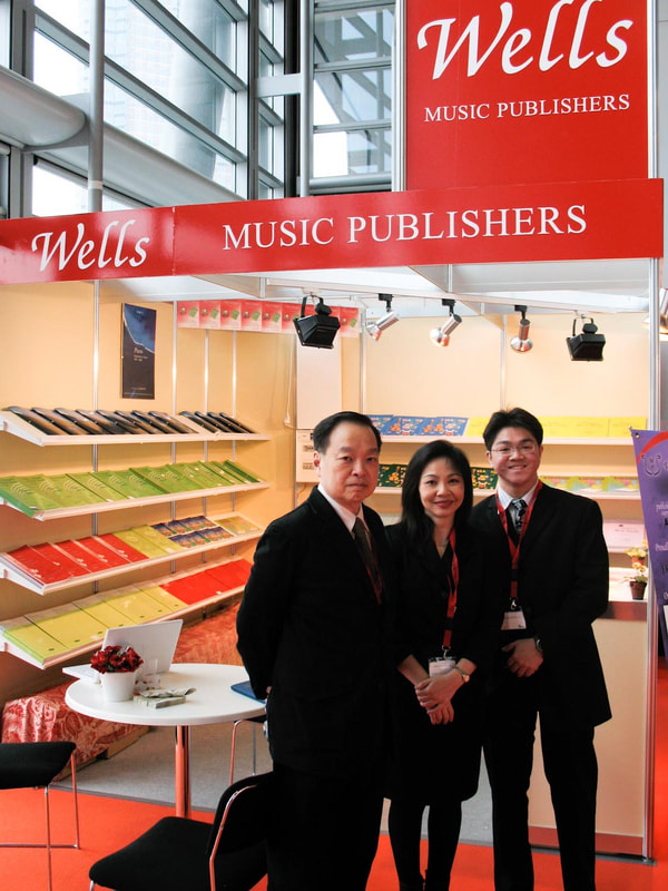 Three people standing inside a Wells Music Publishers booth at a trade show or event, with shelves filled with books or music scores in the background.