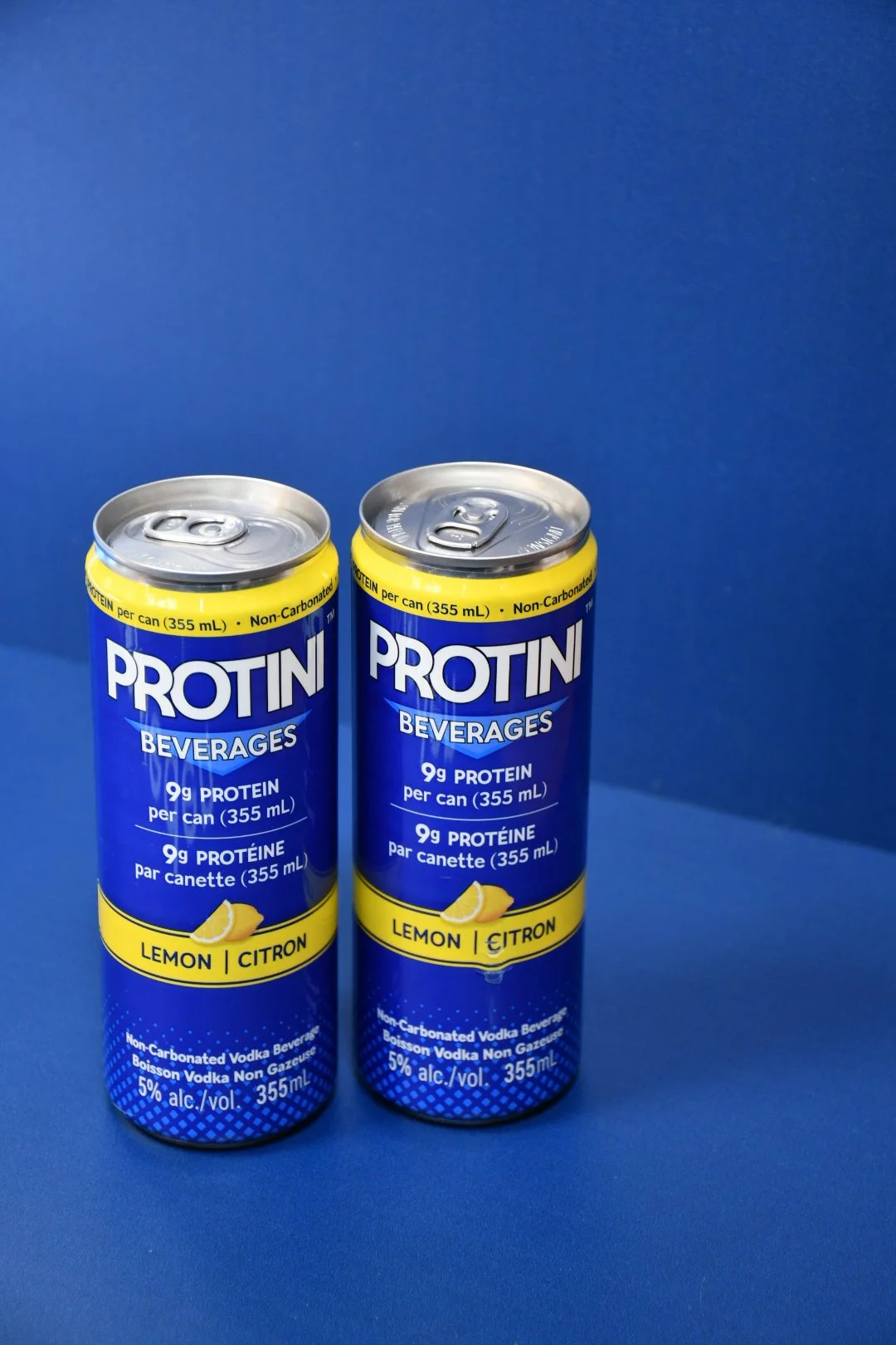 Two cans of Protini lemon flavored beverage and three lemons on a yellow and white striped towel.