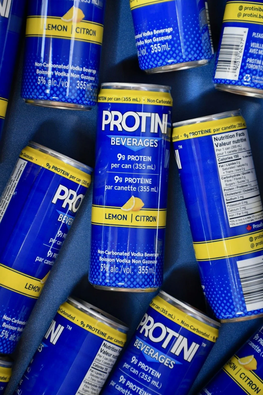 A can of protini vodka beverage with lemon flavour next to a glass of clear water with lemon slices on a white tray.