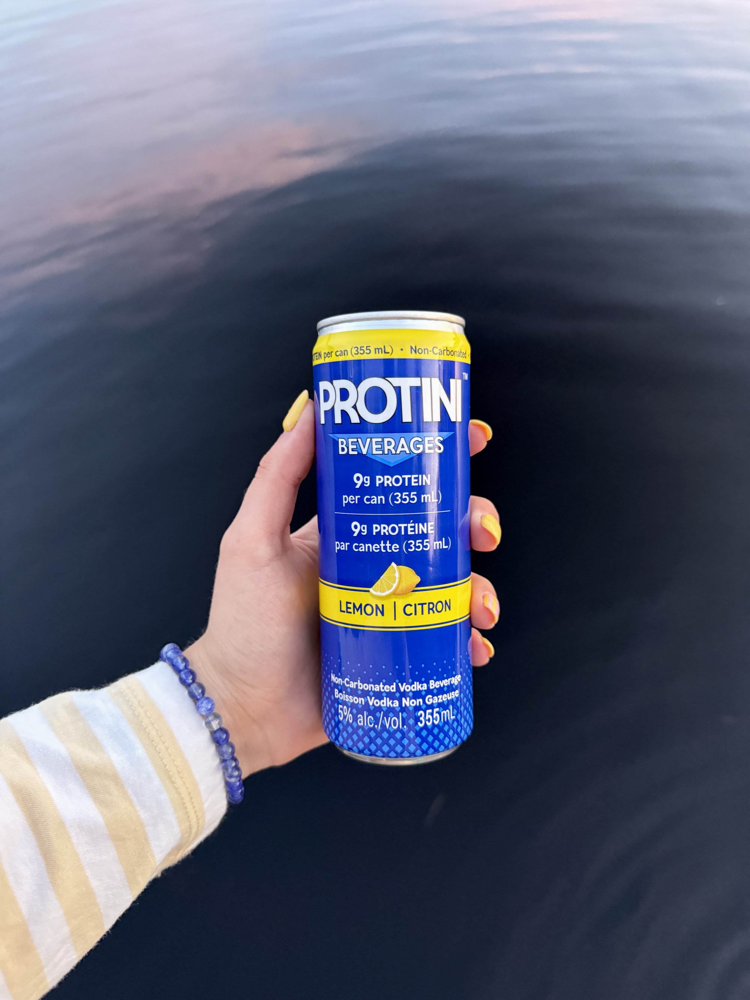 A can of Protini vodka beverage with lemon flavor, placed on a poolside surface next to a pair of sunglasses.
