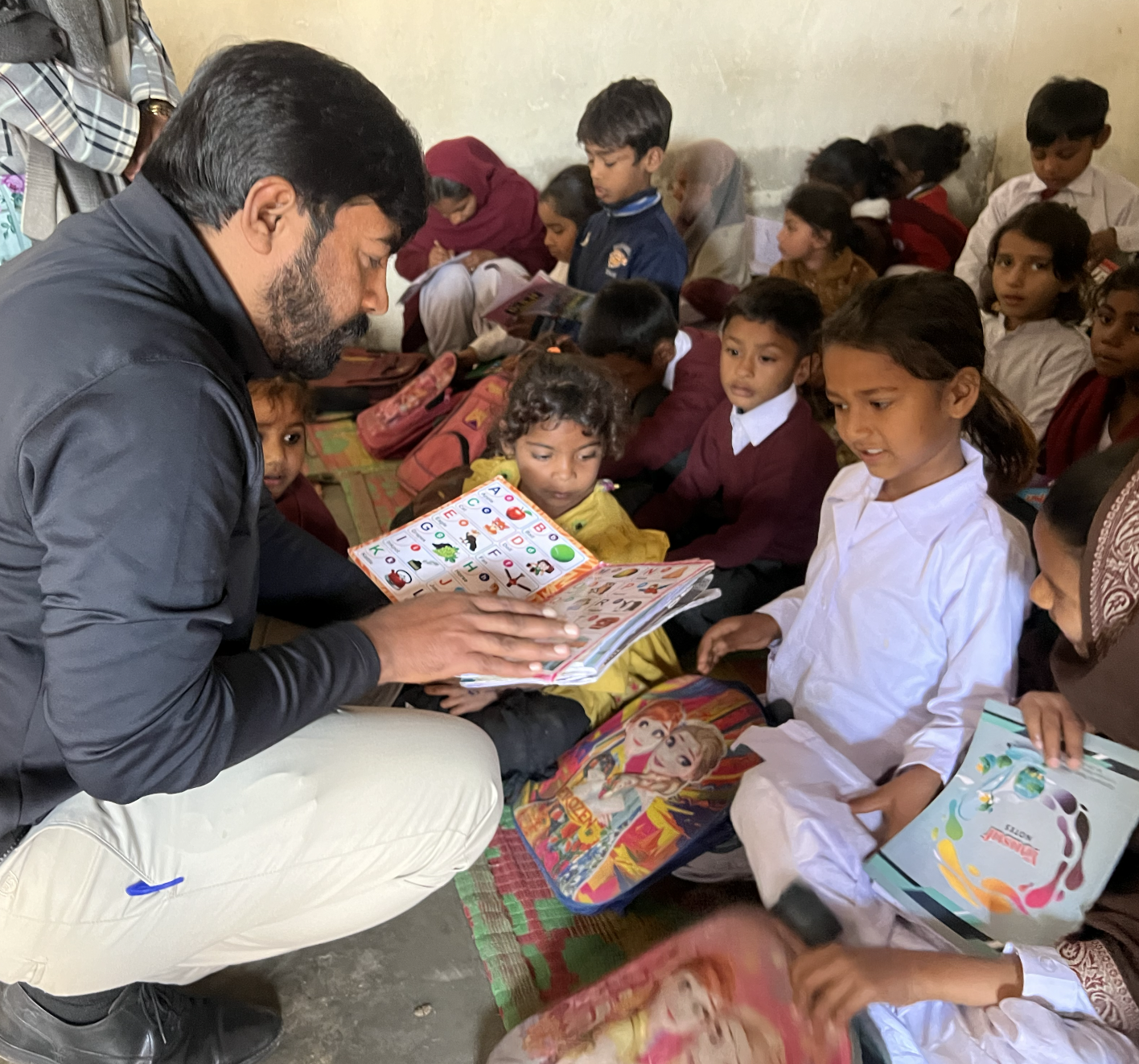 A man with dark hair and beard shows a colorful children's picture book to a group of diverse school children sitting on the floor in a classroom.